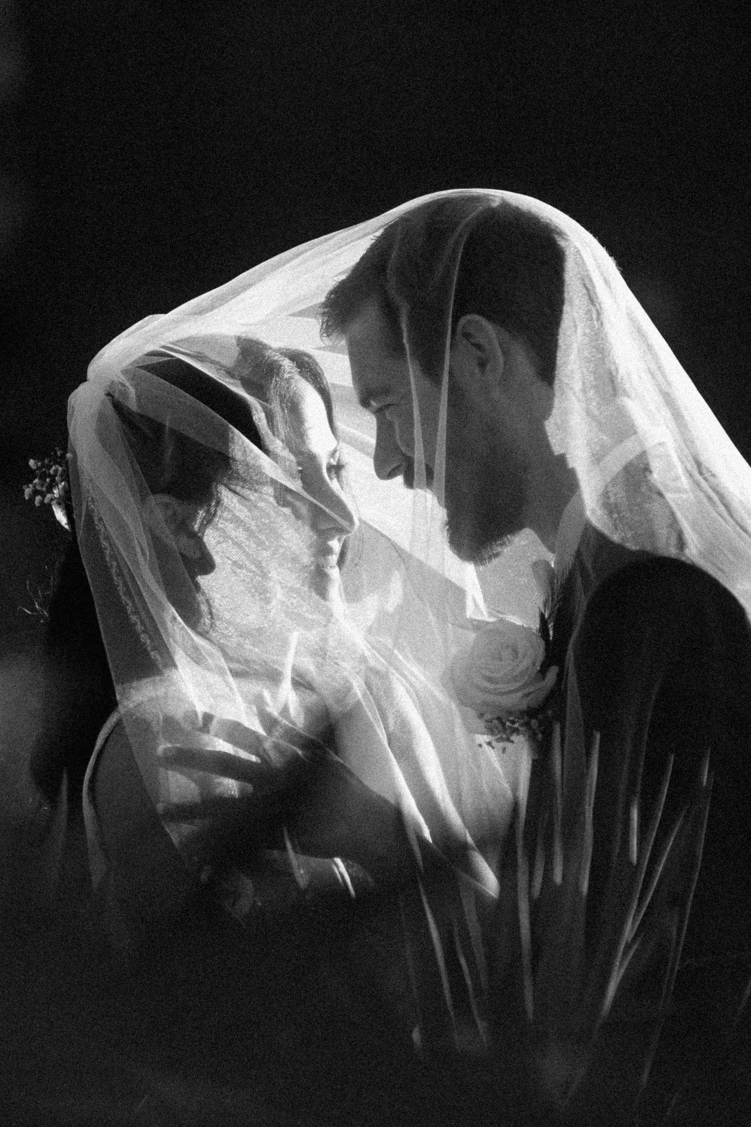 A romantic black and white portrait of a couple sharing a moment under a wedding veil.
