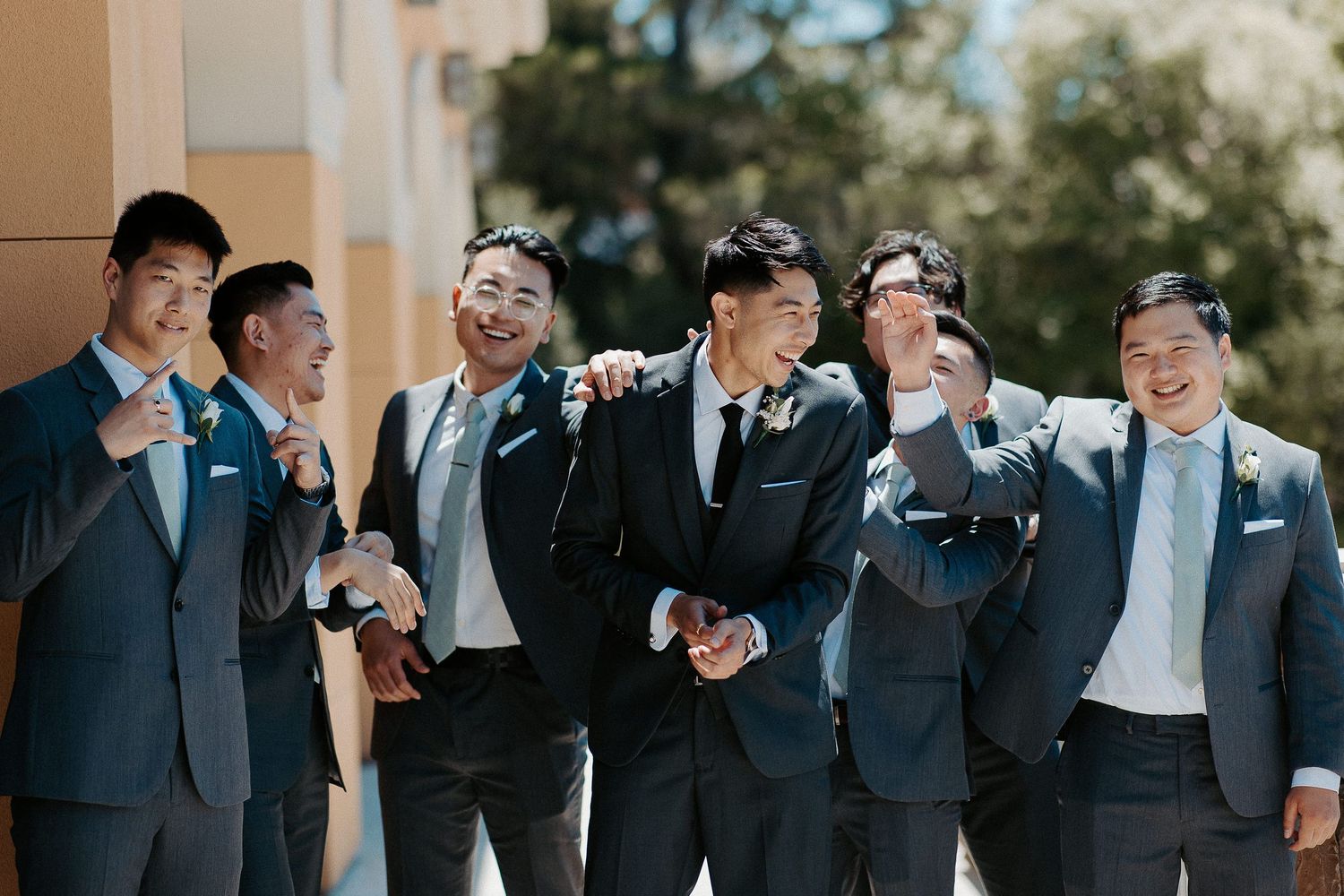 Groomsmen in coordinated navy and gray suits share a candid laughing moment together outdoors.