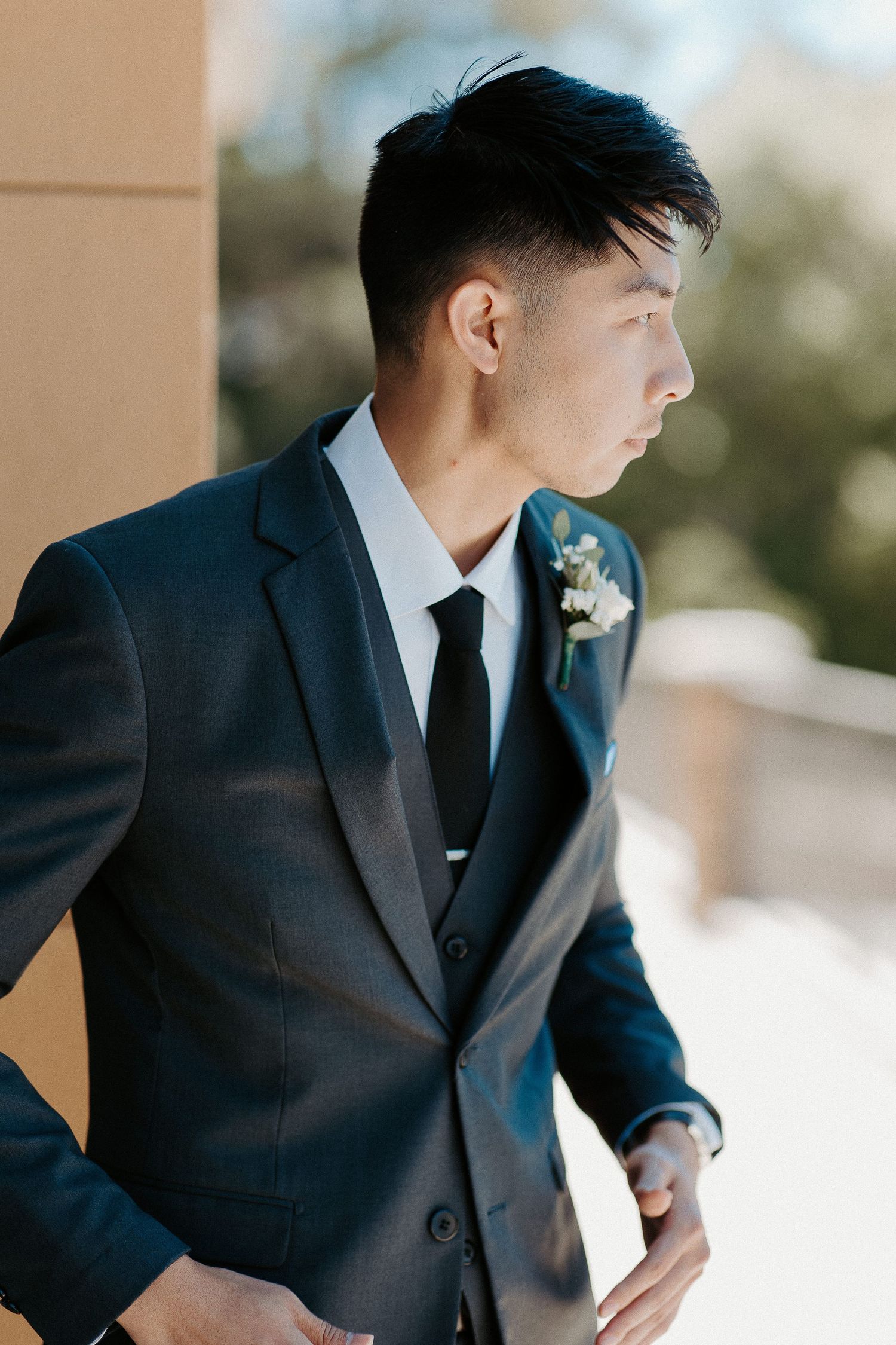 Side profile of groom wearing dark suit with boutonniere looking away outdoors in natural lighting.