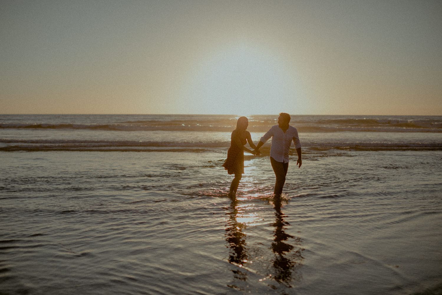 Couple holding hands while standing in shallow ocean water at sunset.