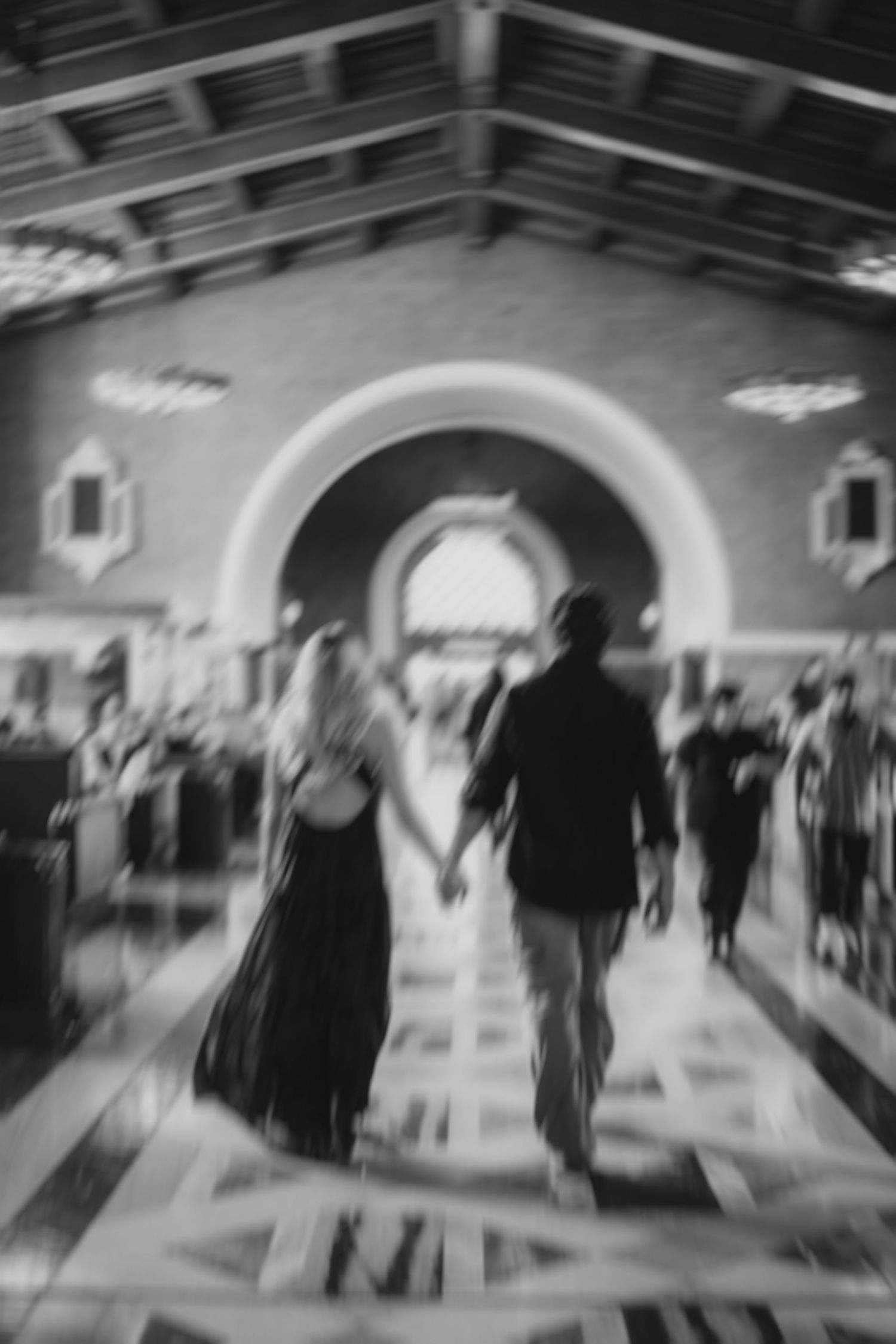 A blurred black and white image of figures walking through an arched corridor.