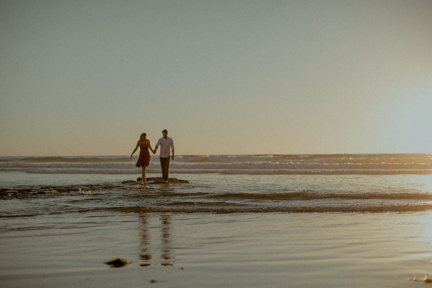 Two figures walking hand in hand through shallow ocean water at golden hour.