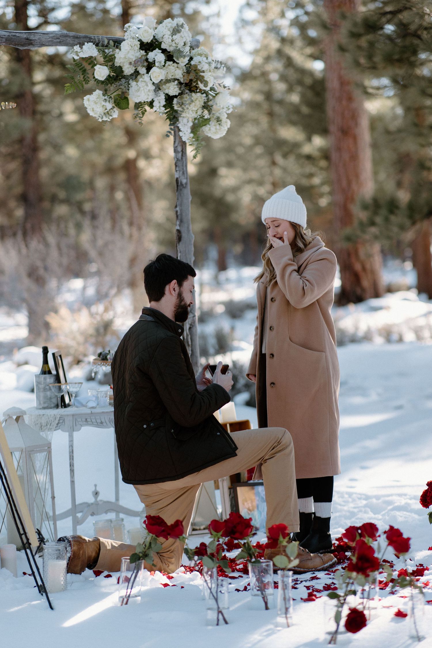 A winter proposal moment beneath white floral decorations with red rose petals in the snow.