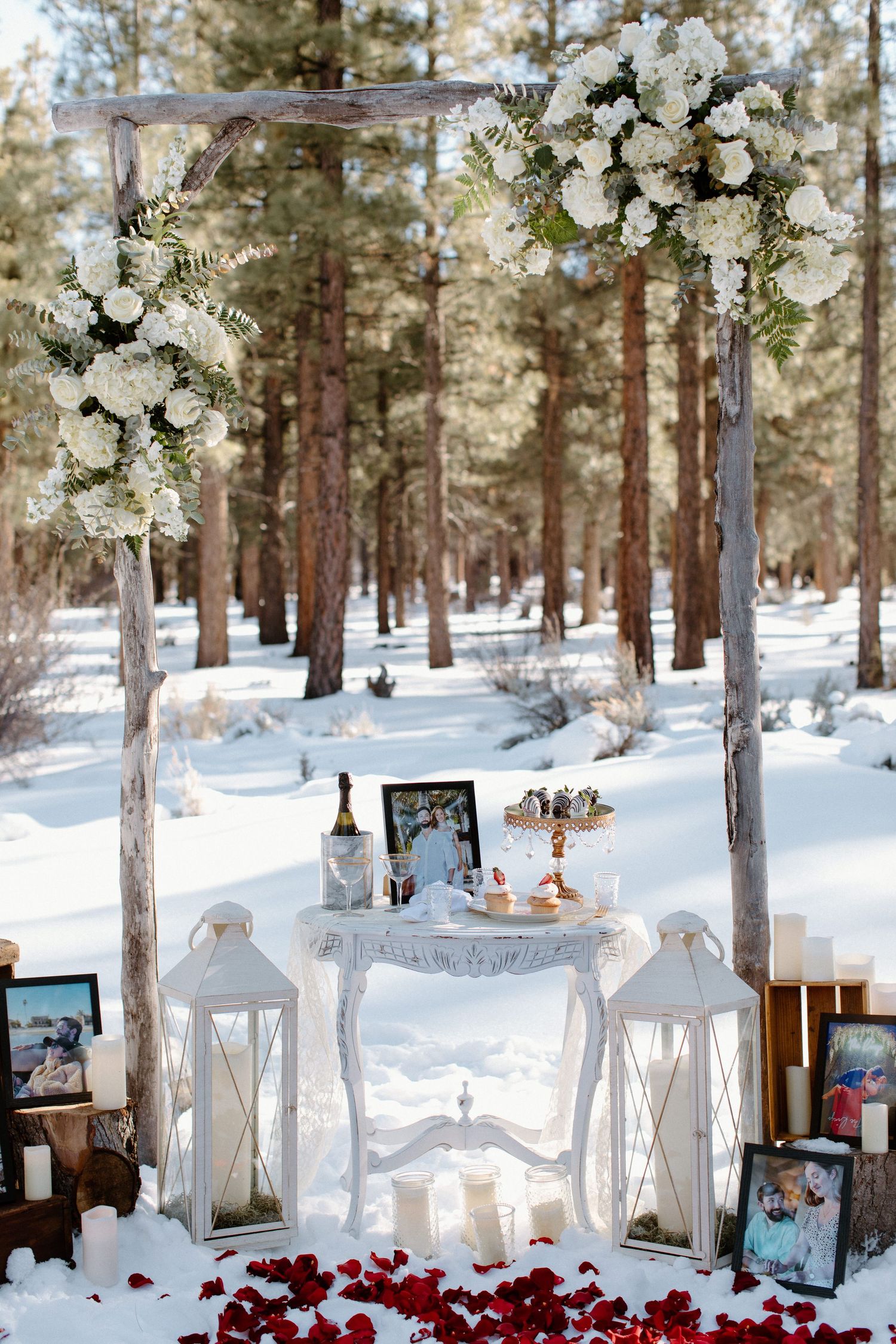 White floral arch with lanterns and decorative table setup in snowy forest setting.