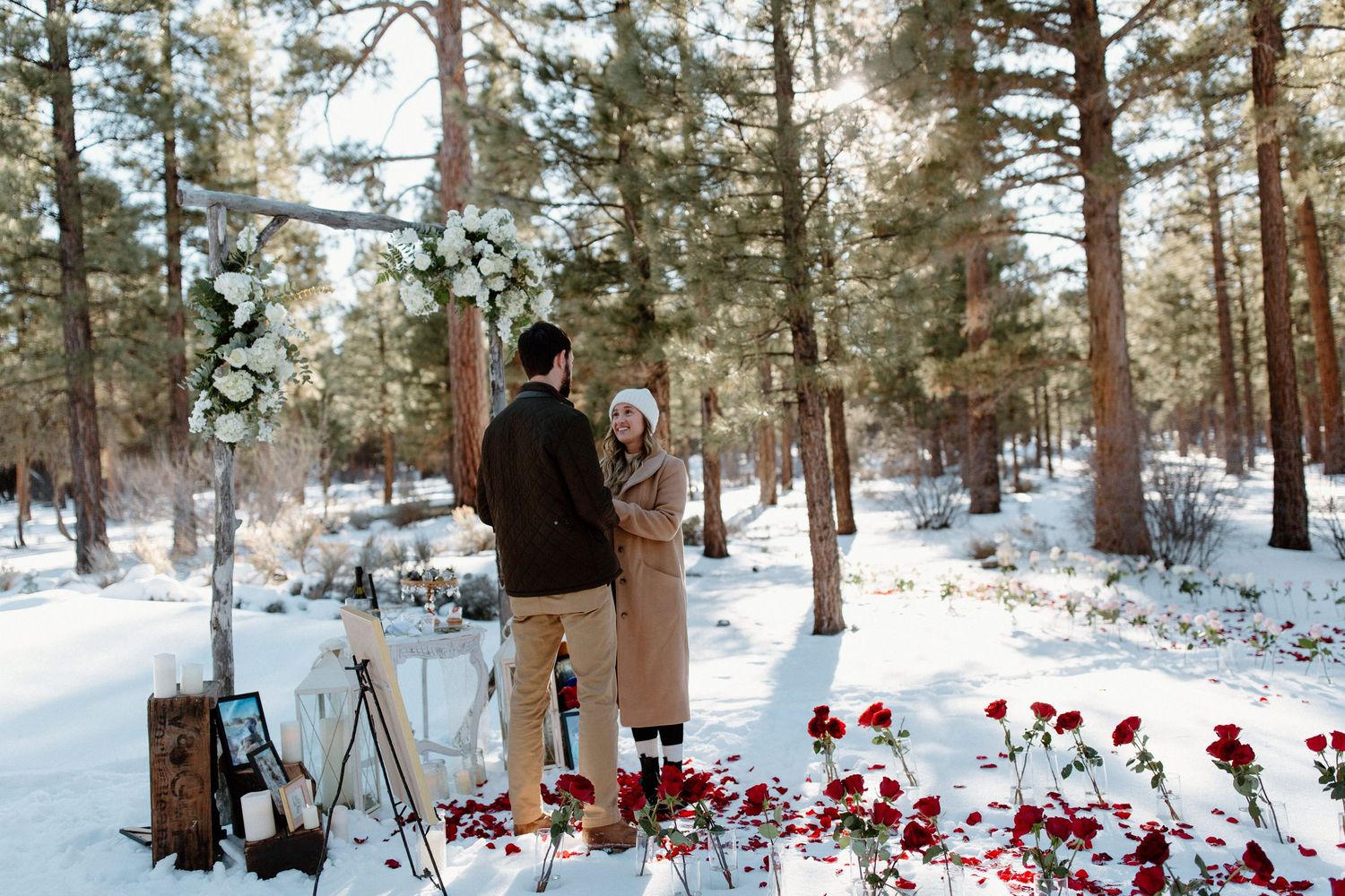 Couple standing under decorated floral arch in snowy woods with red roses scattered around.