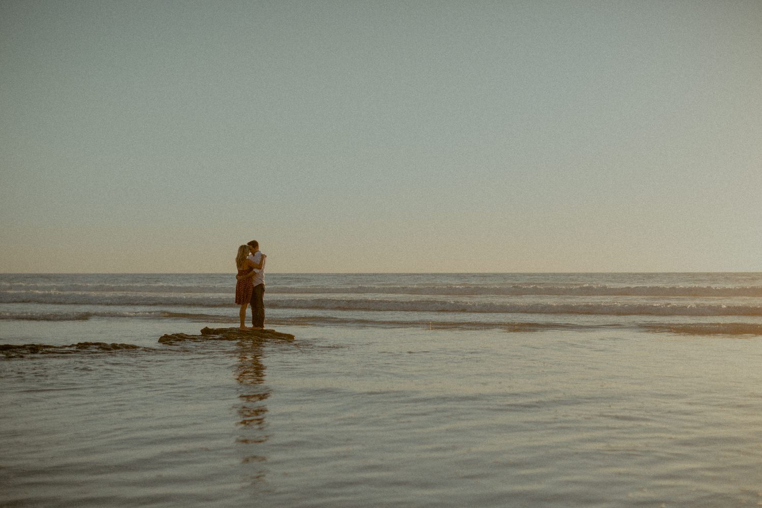 A silhouetted couple stands together at the edge of the ocean during sunset.