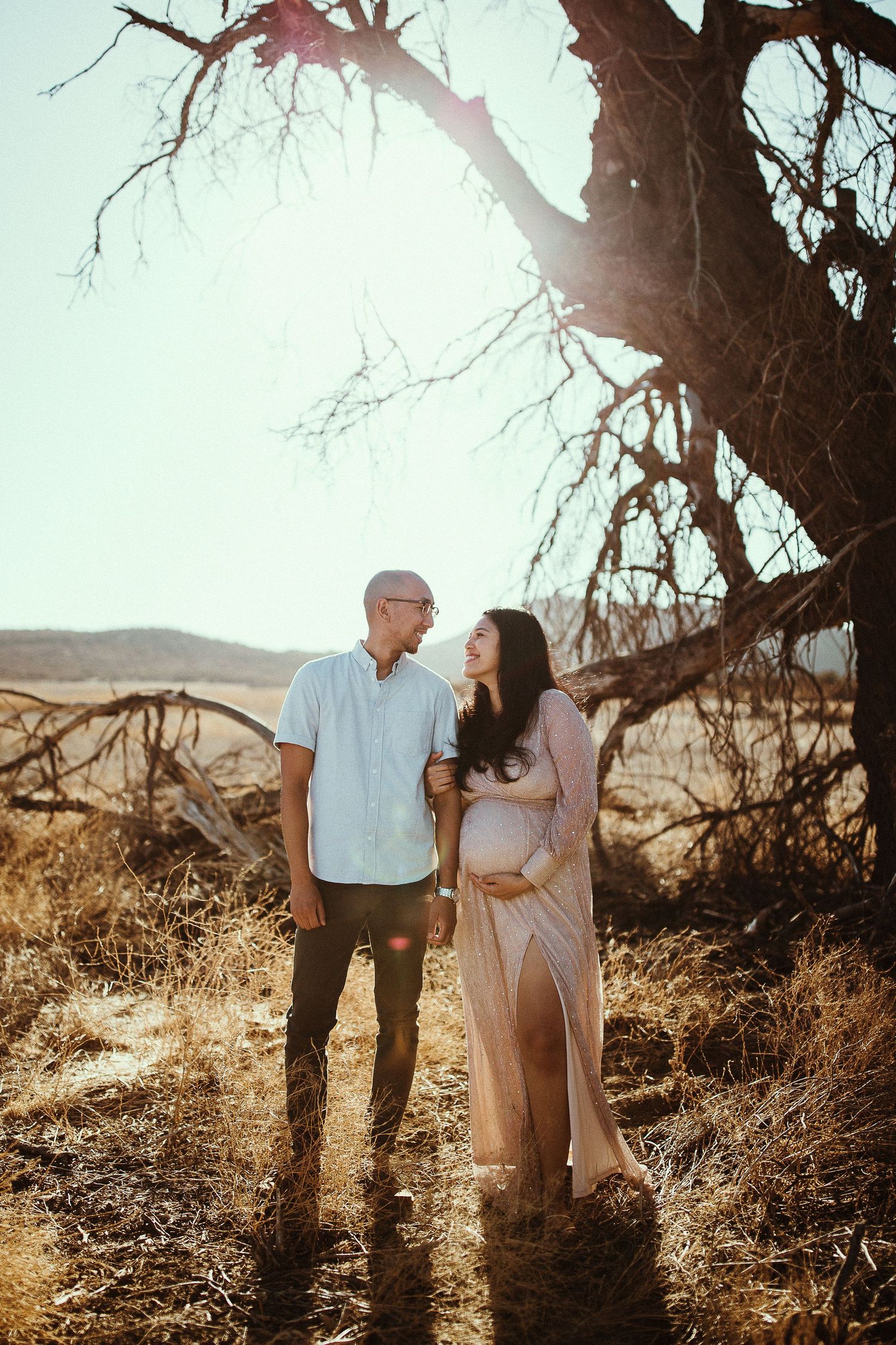 Romantic maternity photo of couple standing under tree branches in desert setting at sunset.