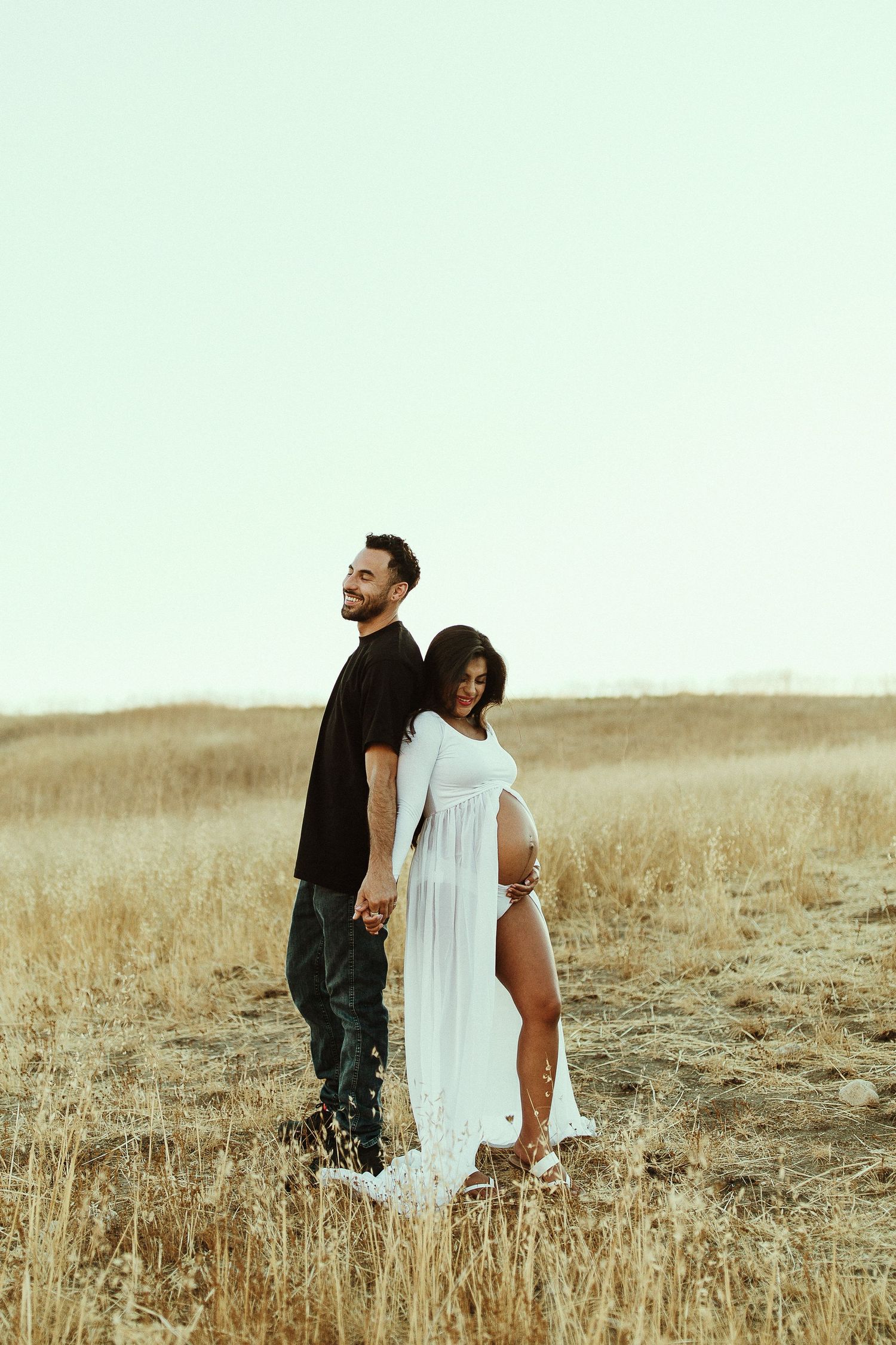 Artistic maternity portrait of couple standing back-to-back in wheat field.
