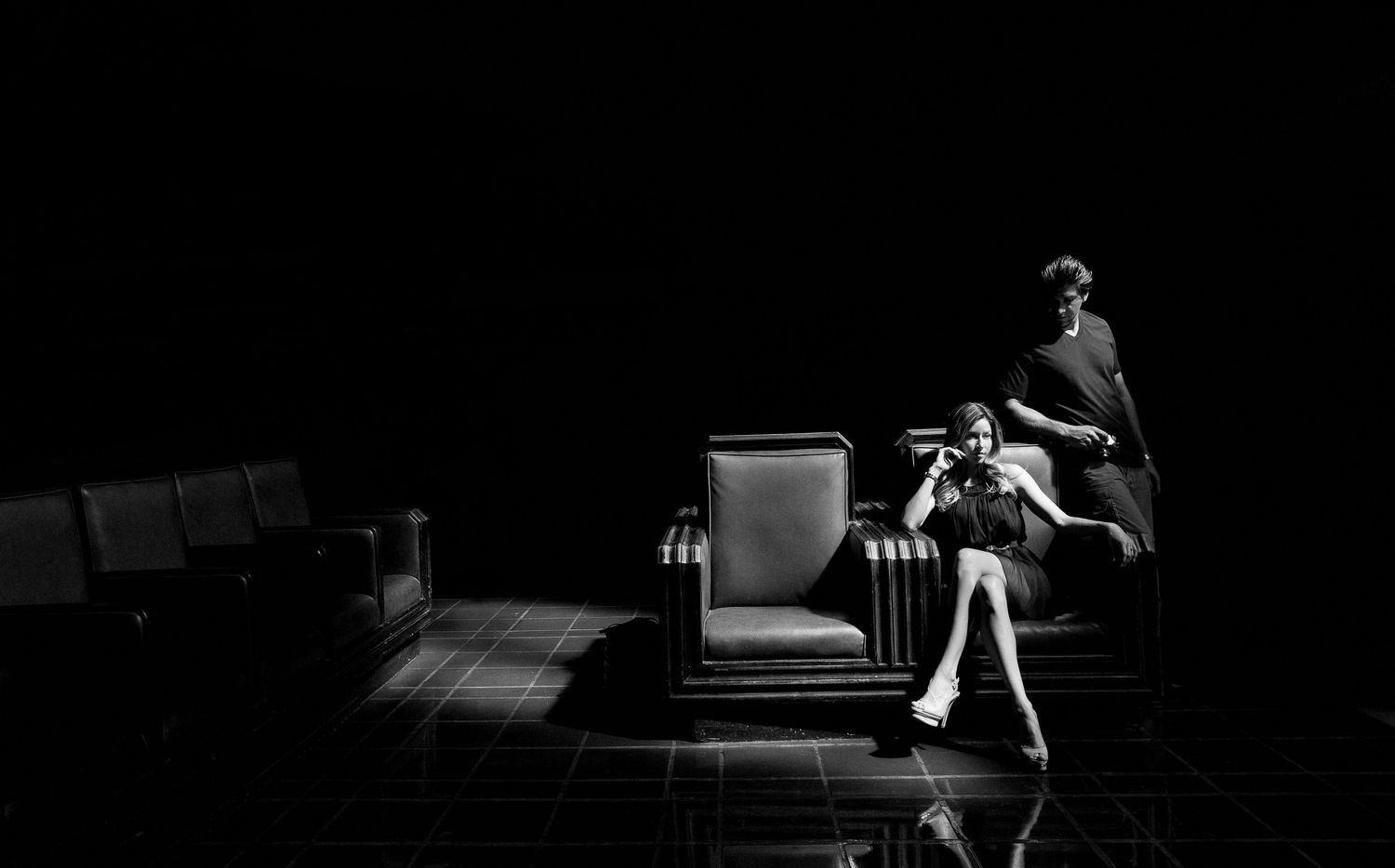Dramatic black and white theater stage scene with a single figure seated in a spotlight.