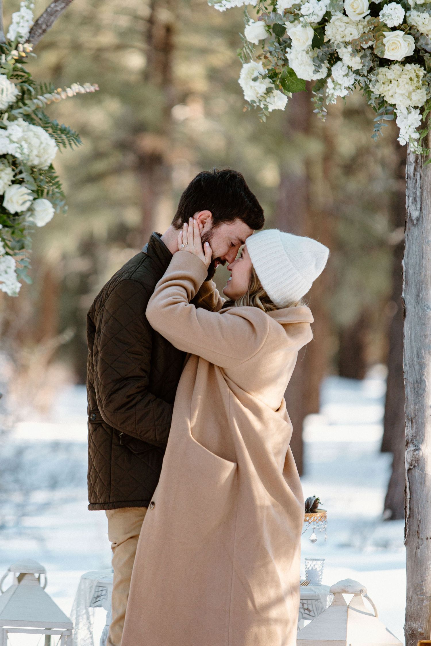 A romantic embrace under white floral decorations in a snow-covered forest setting.