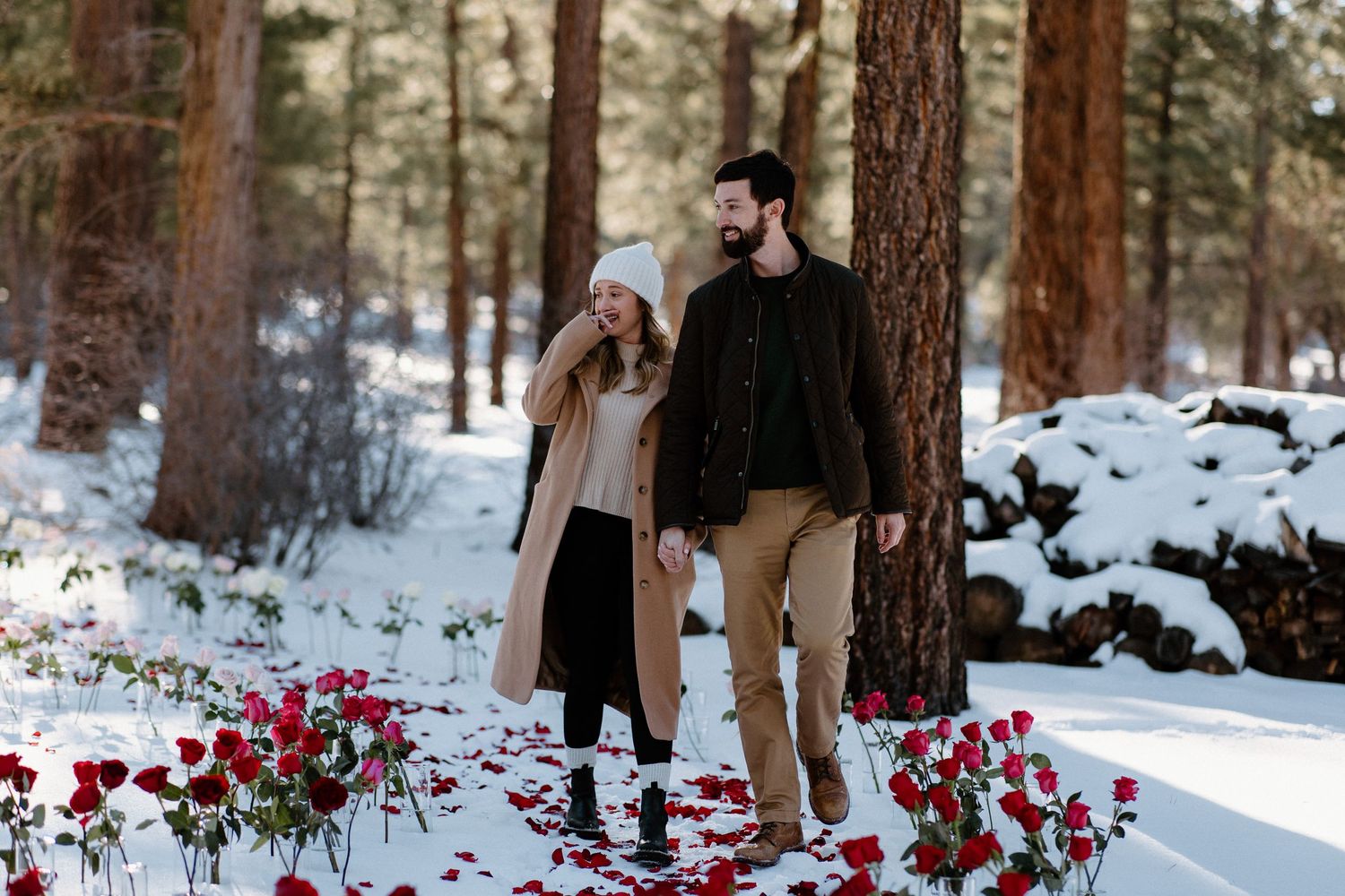 A couple walks through a snowy forest with red rose petals scattered on the ground beneath pine trees.