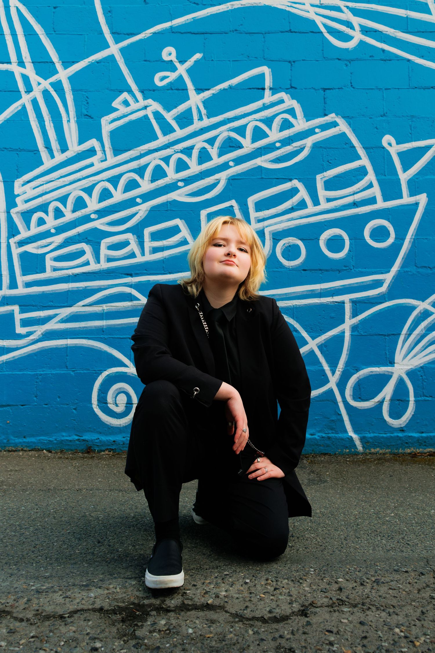 A person in black attire crouches against a bright blue wall with white ship mural artwork.