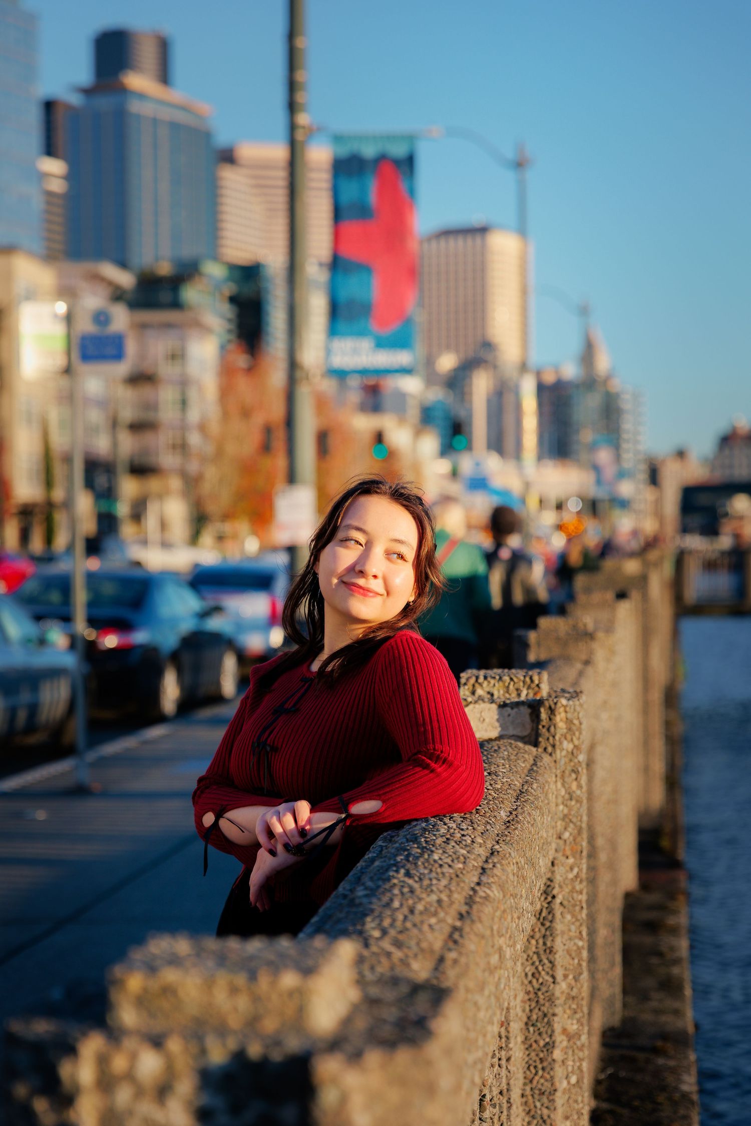 Waterfront scene showing a girl in a red sweater leaning on a stone wall with a city skyline in the background.