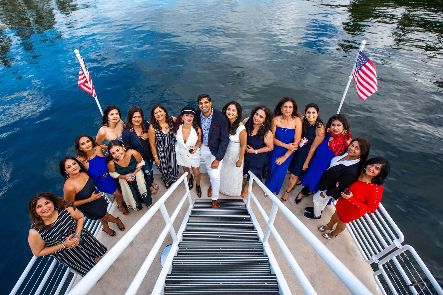 Large group in formal attire poses on white dock stairs with American flags and water backdrop.