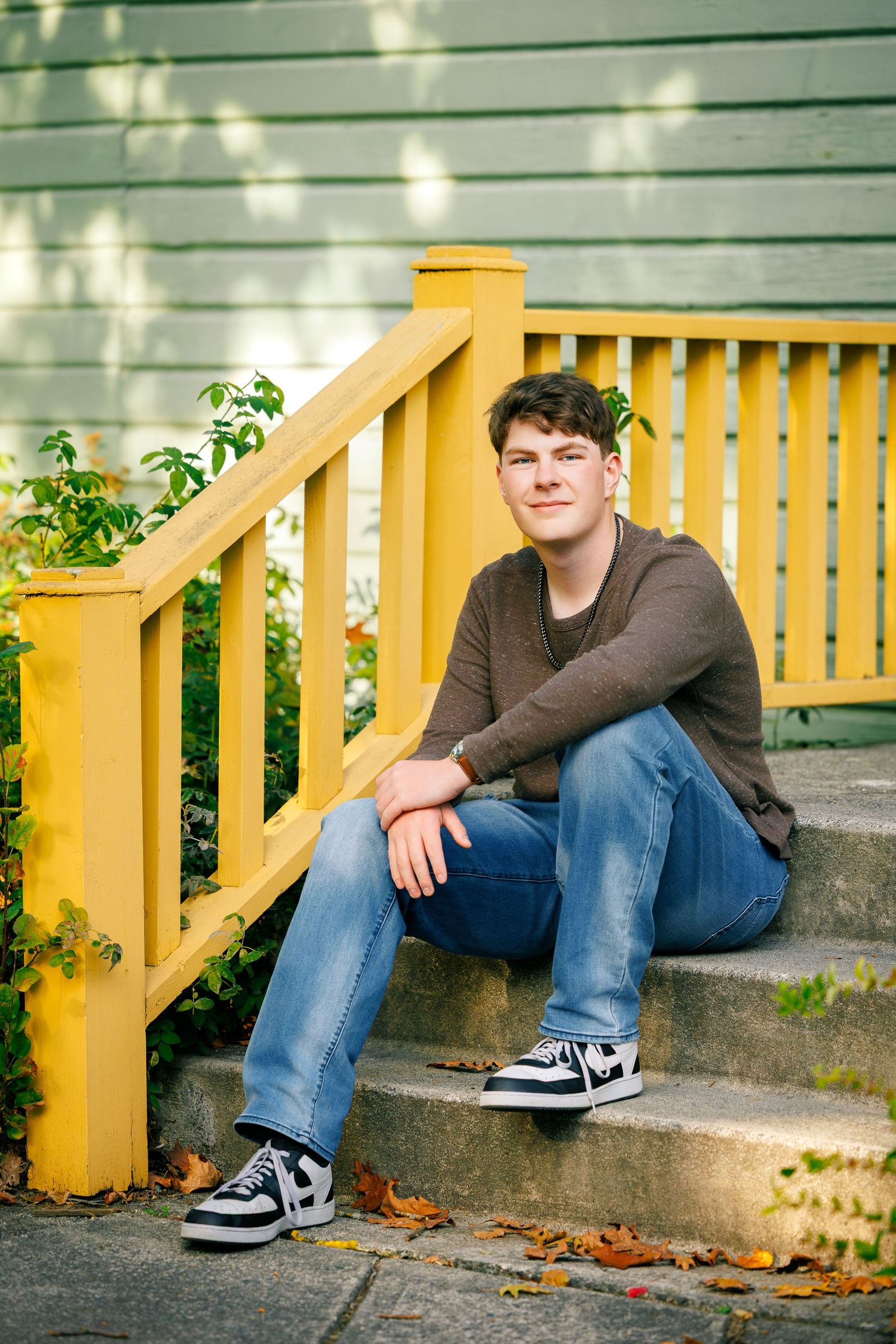 Someone sits casually on outdoor concrete steps with a yellow painted railing, wearing jeans and a brown sweater.
