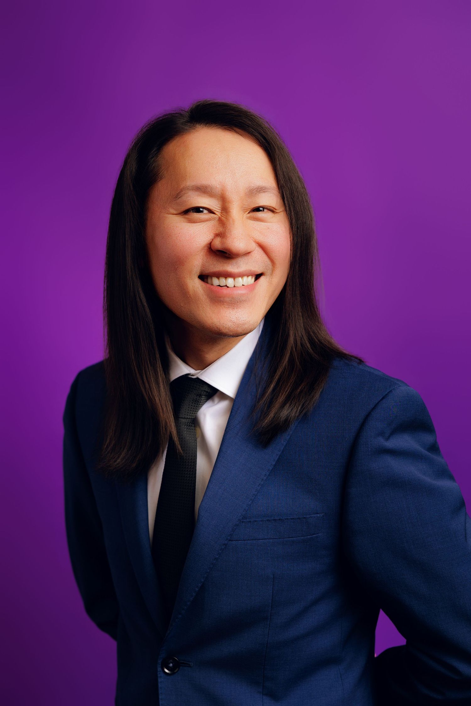 Professional headshot of someone in a navy blue suit and black tie smiling against a vibrant purple background.