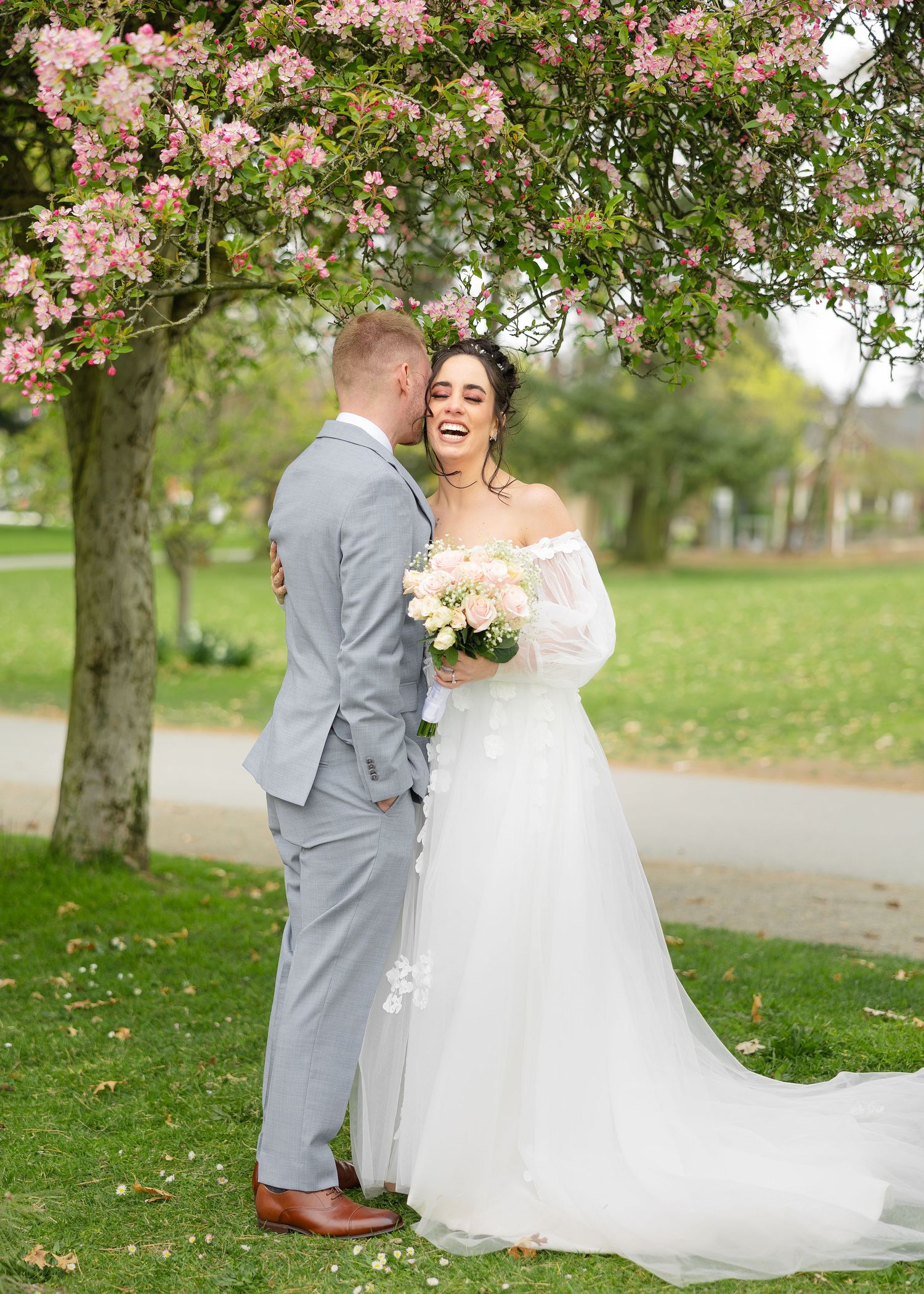 A couple walks under blooming cherry blossom trees on a spring day along a garden path in their wedding attire.