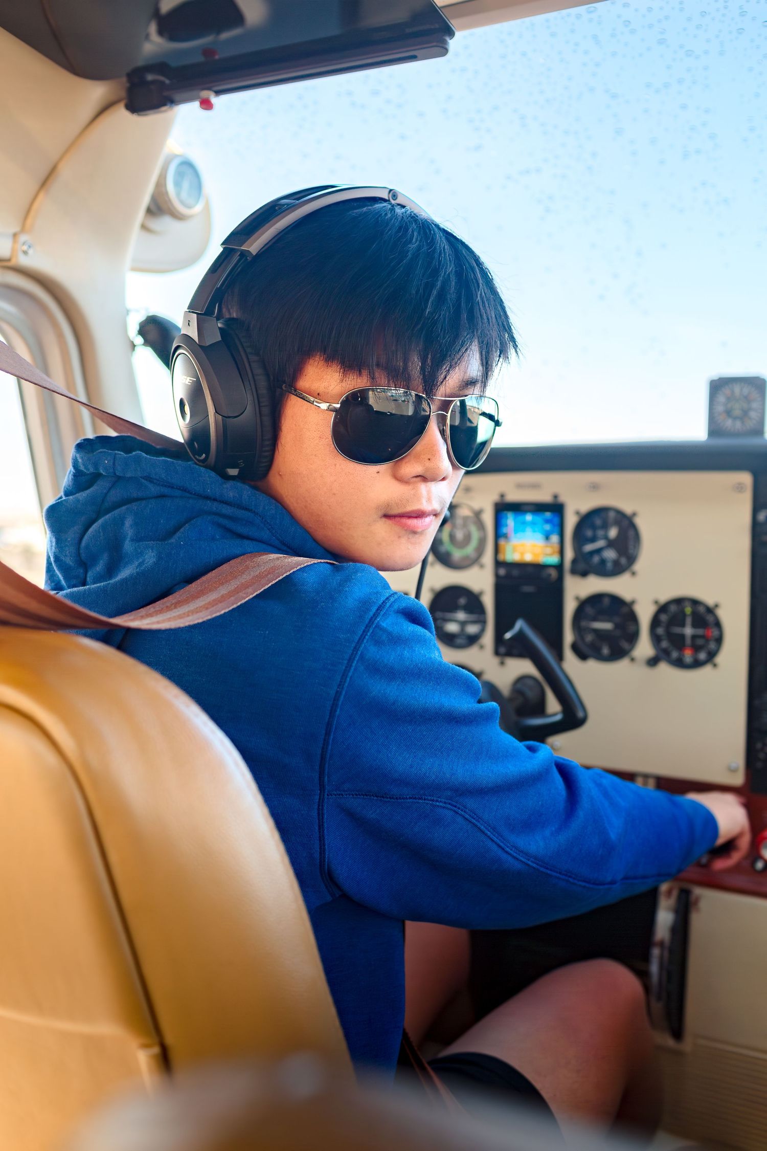 A person wearing sunglasses and headphones sits in the cockpit of a small aircraft with tan leather seats.