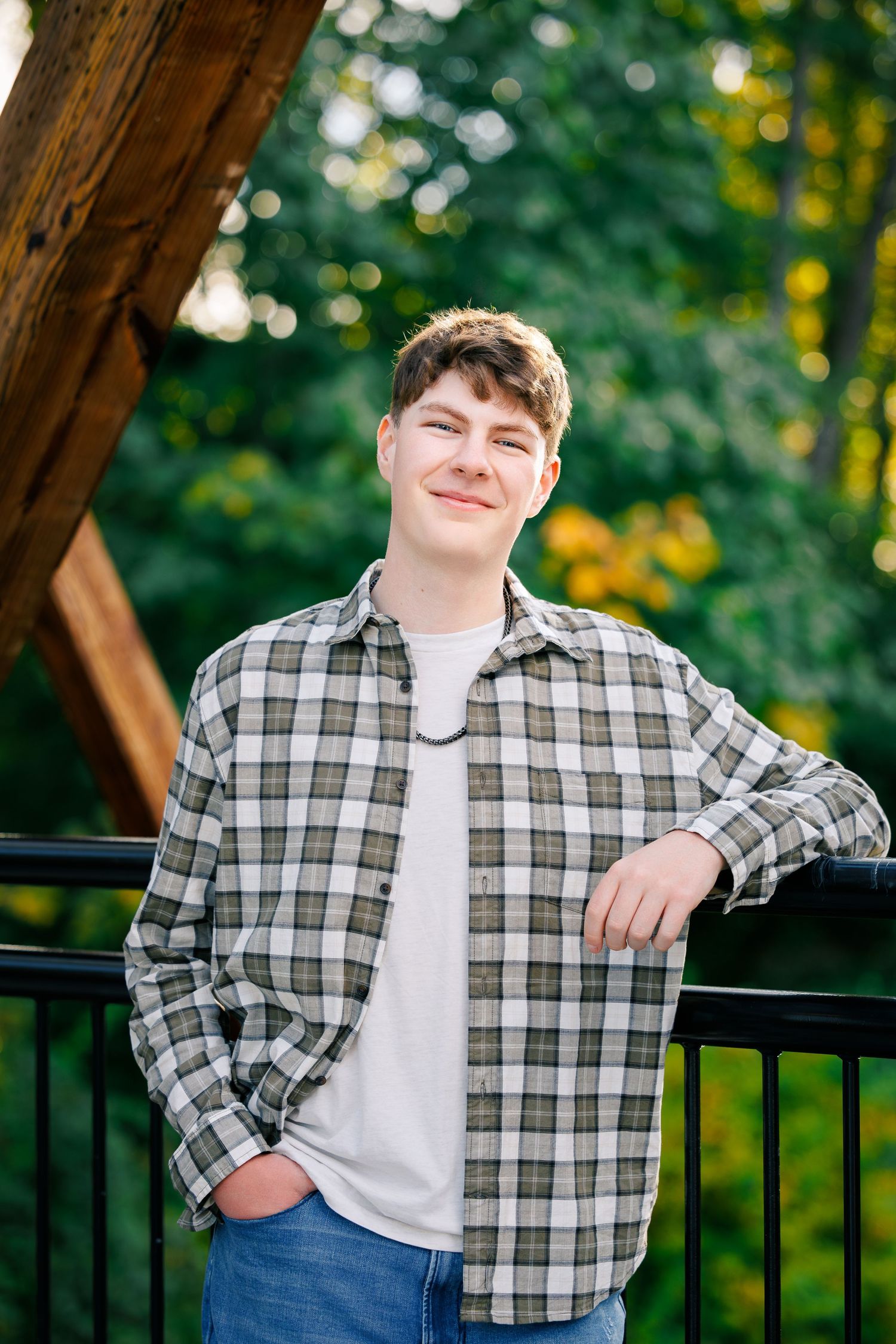 A person in a plaid flannel shirt and white t-shirt poses casually against a railing with trees in the background.
