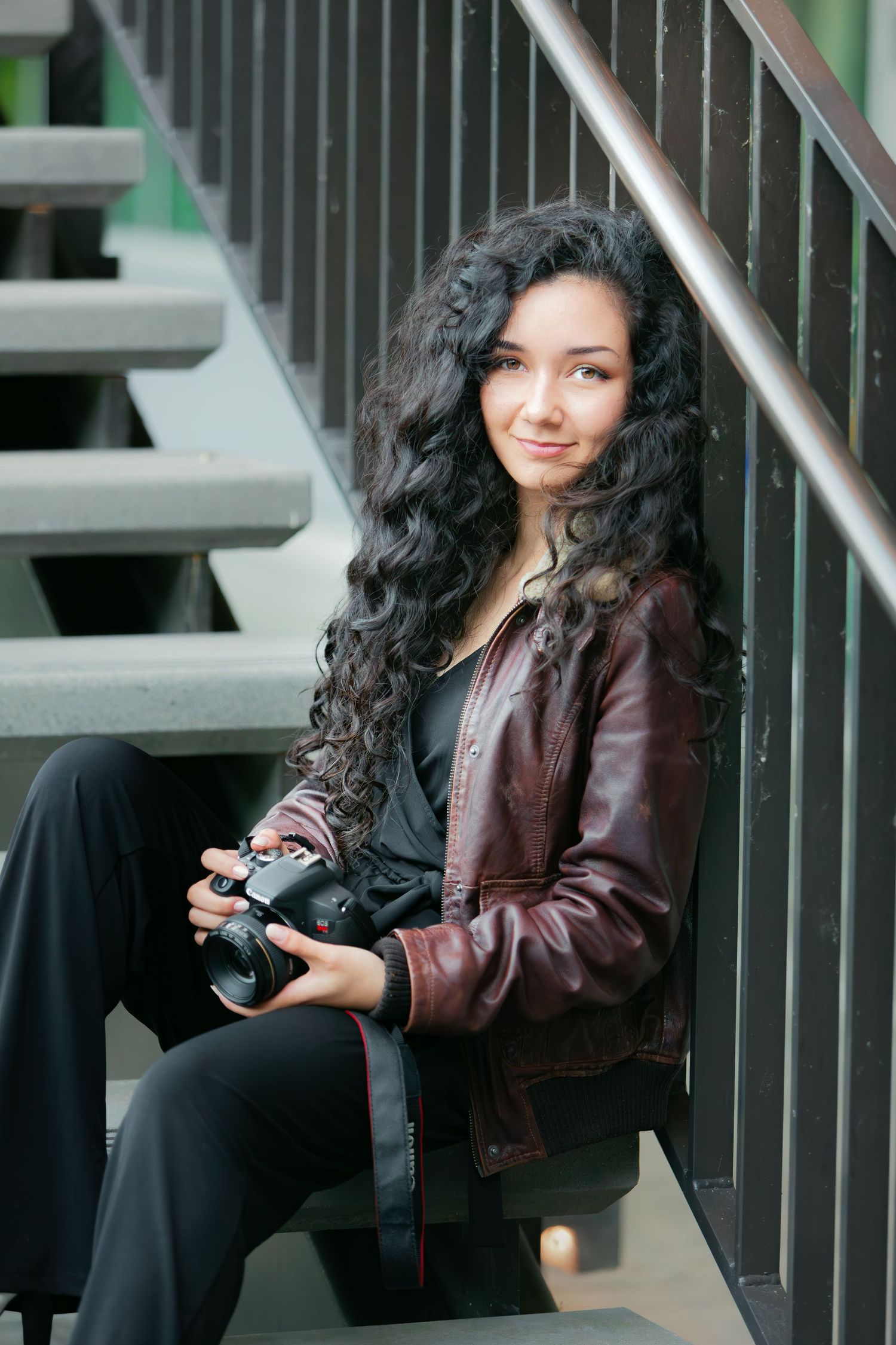 A curly haired senior girl sits on concrete steps holding her camera