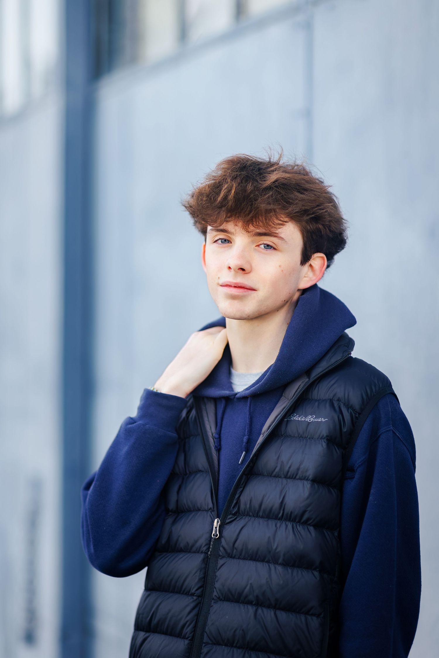 Person in navy hoodie and black puffer vest poses thoughtfully against modern architectural backdrop.