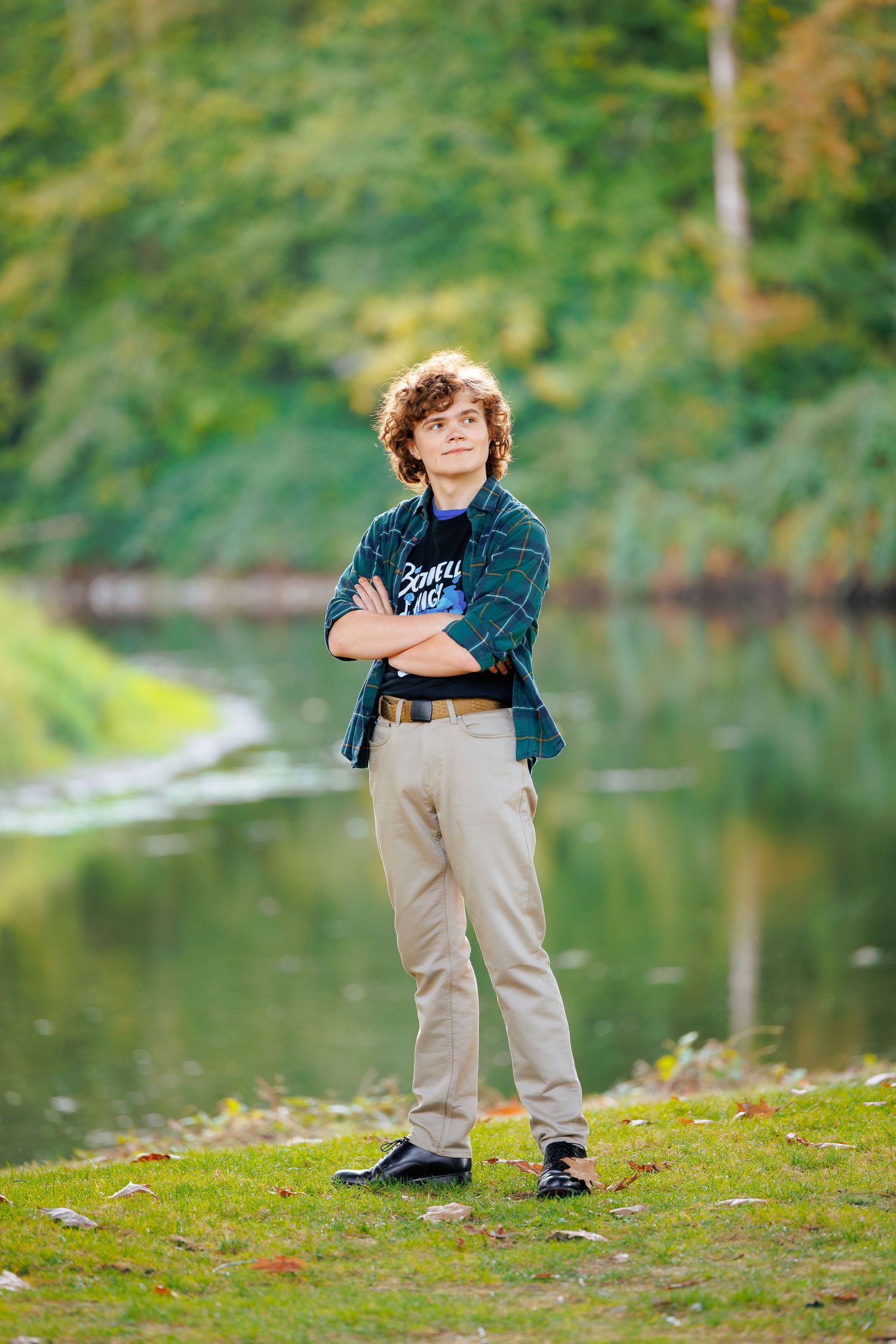 A child wearing a plaid shirt and khaki pants stands on a grassy bank beside a tranquil lake on an autumn day.