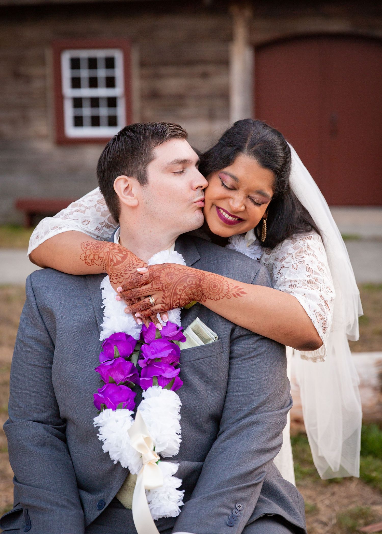 Two newlyweds share a tender moment outside a rustic barn with purple and white flowers and gray suit details.