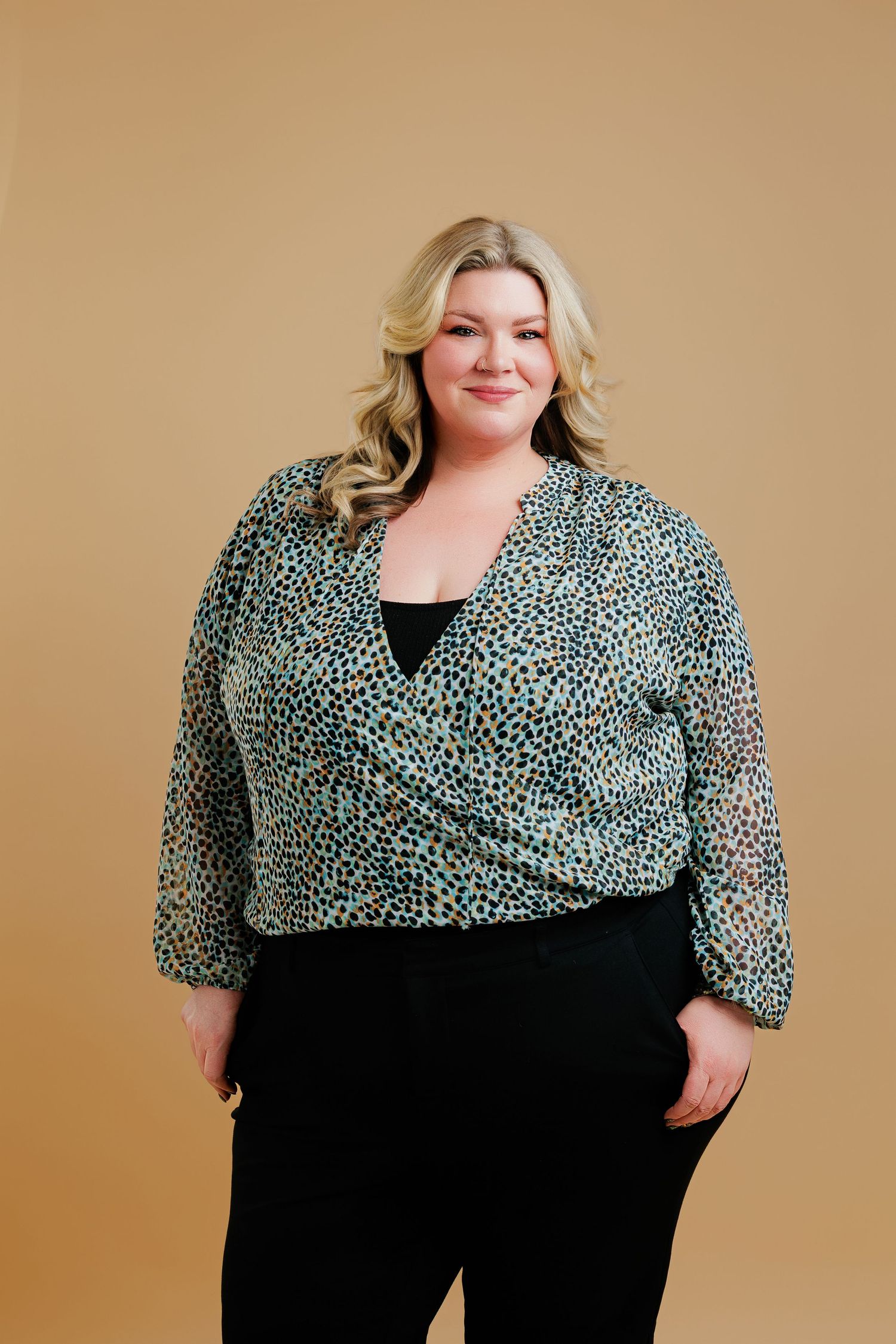 Plus size model wearing a black and white patterned wrap top and black pants against a beige backdrop.