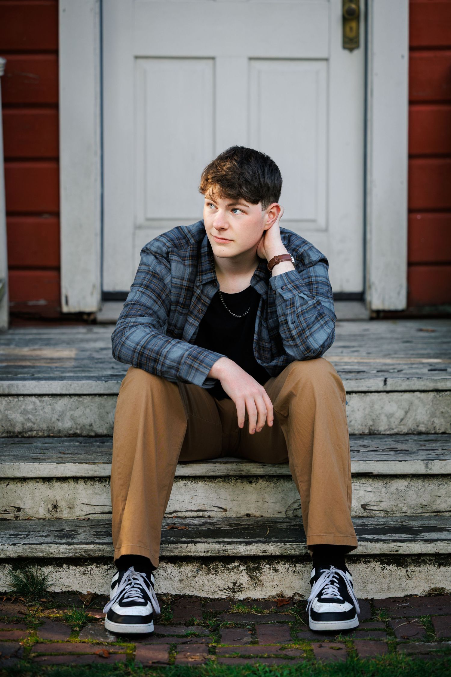 Person wearing plaid flannel and khakis sits on concrete steps in front of red building with white doors.