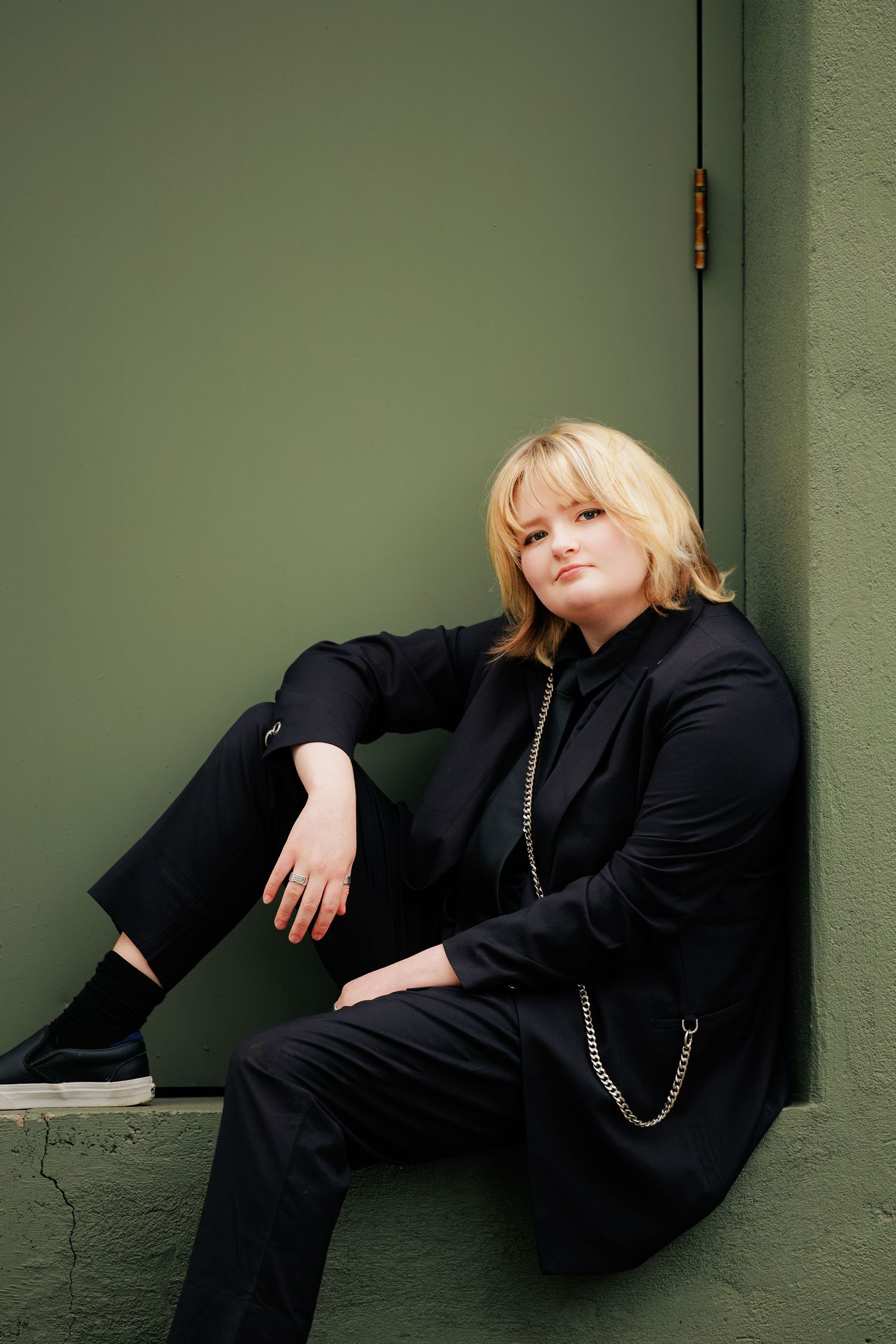 A figure in an all-black outfit sits casually against a green wall with a chain accessory detail.