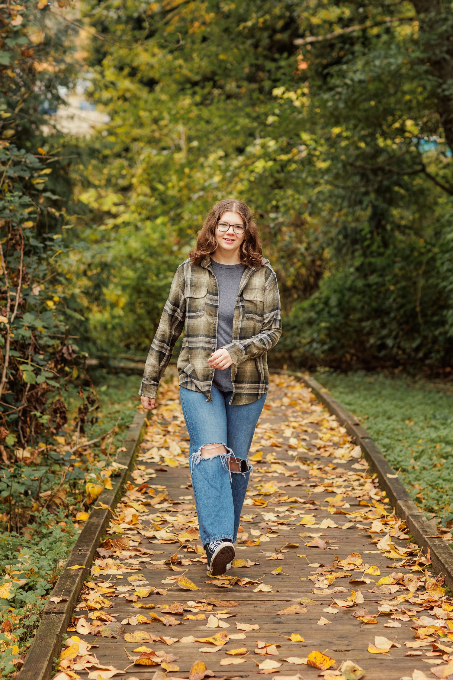 A person walks along a wooden boardwalk path covered in colorful autumn leaves surrounded by lush green foliage.