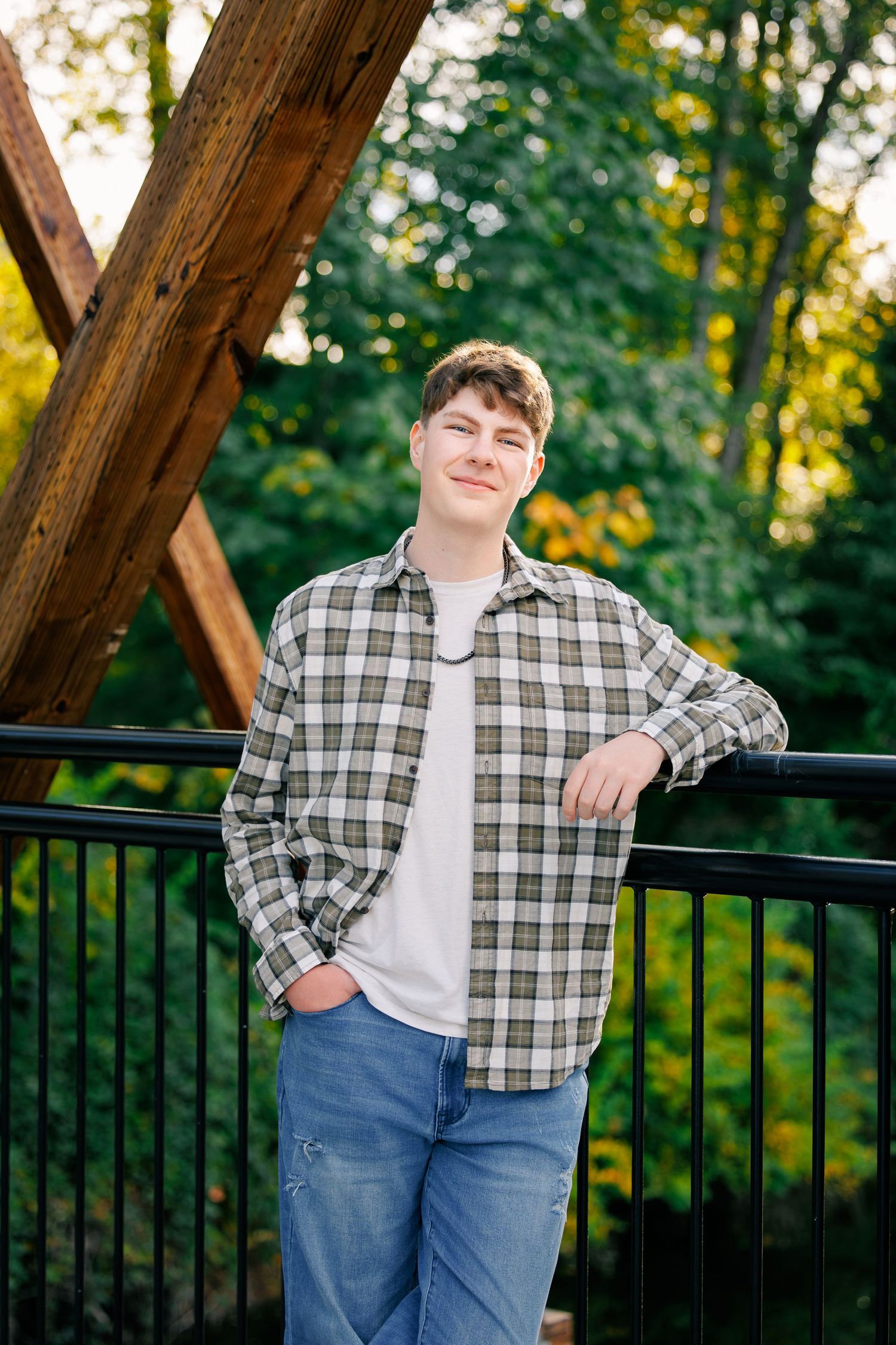 A casual outdoor portrait showing someone in a plaid flannel shirt and jeans leaning against a black railing at sunset.