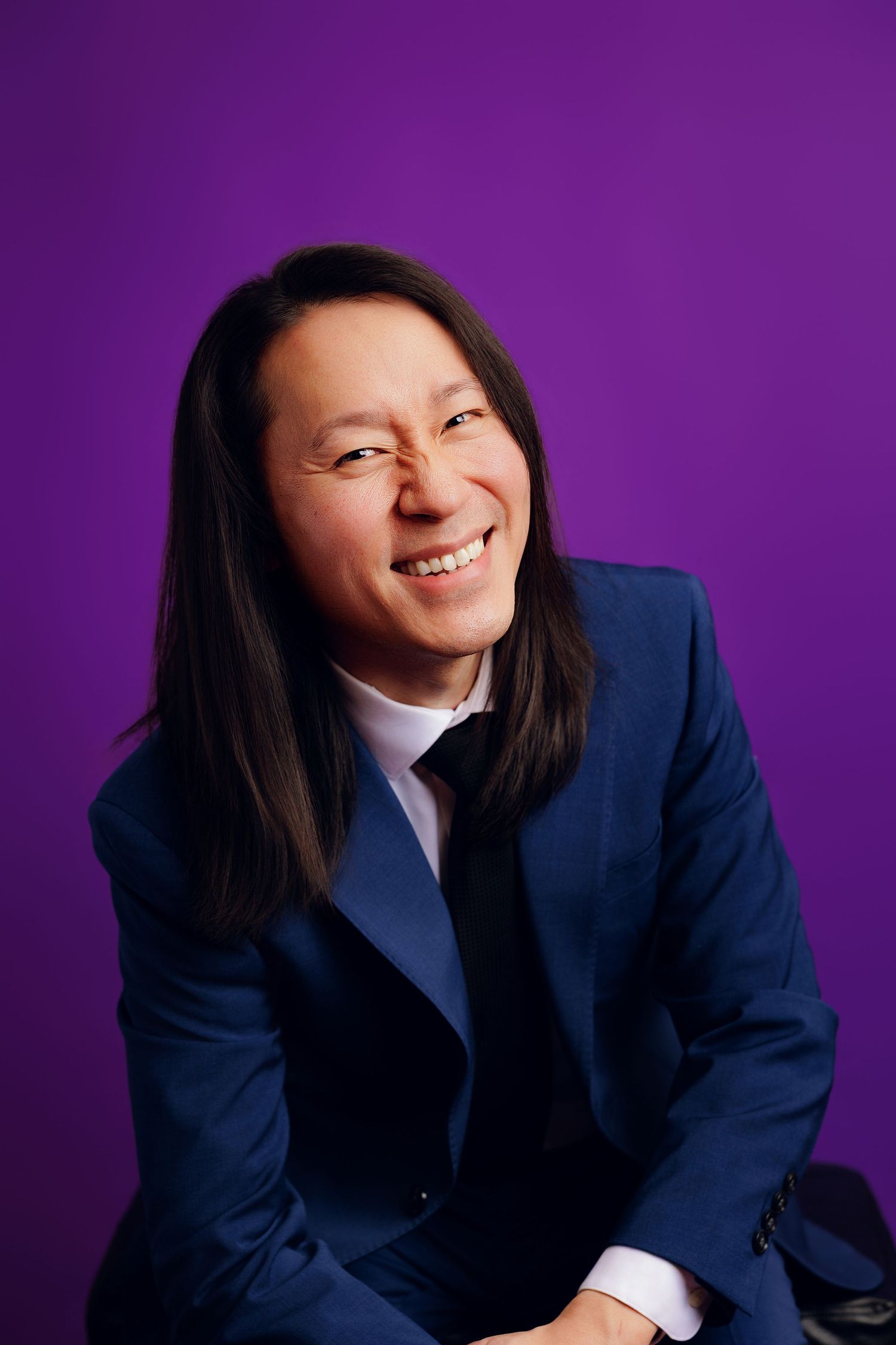 A warm portrait of someone with long dark hair wearing a navy business suit against a vibrant purple background.