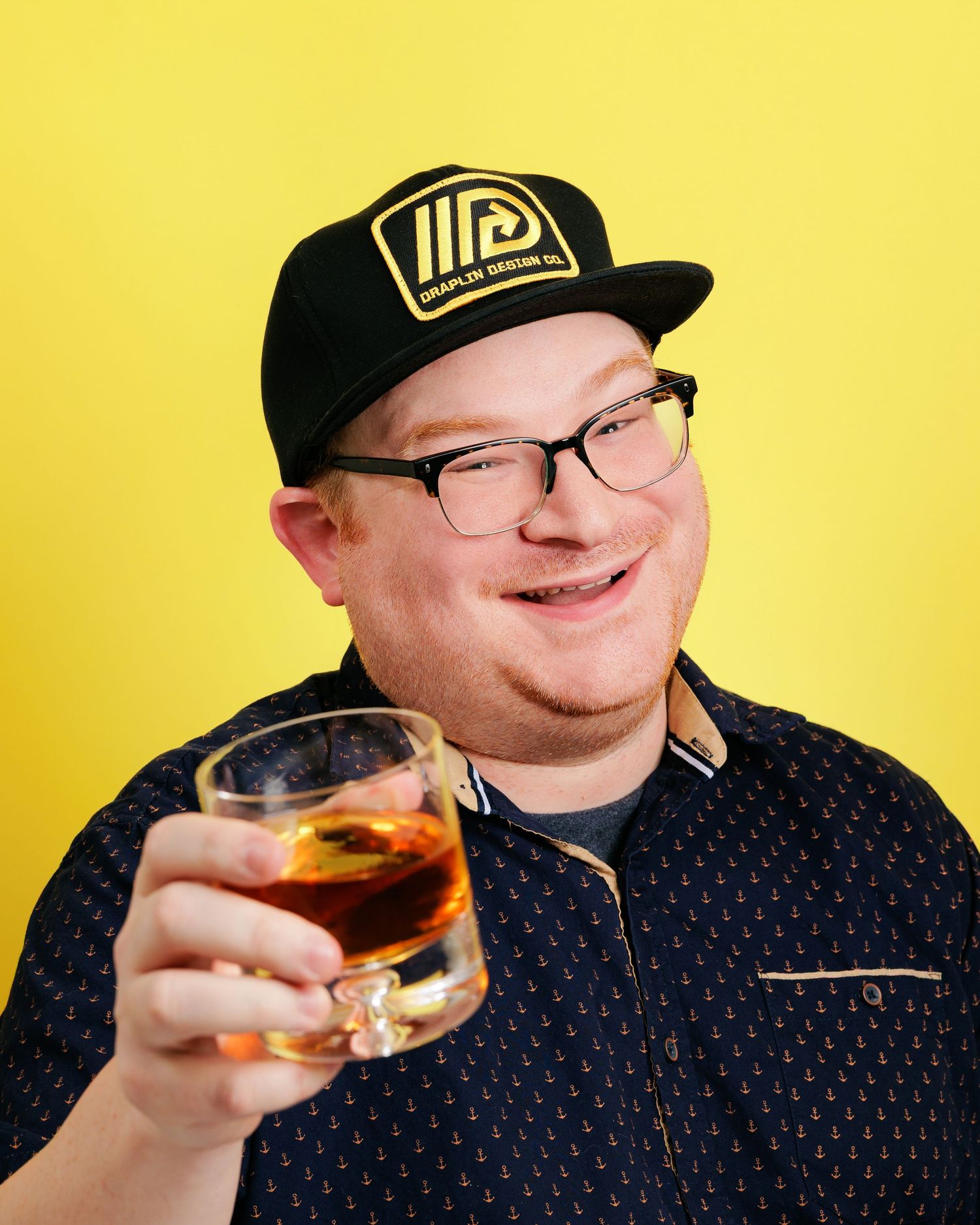 A man wearing glasses and a black baseball cap holds up a glass of dark liquor while sitting against a bright yellow background.