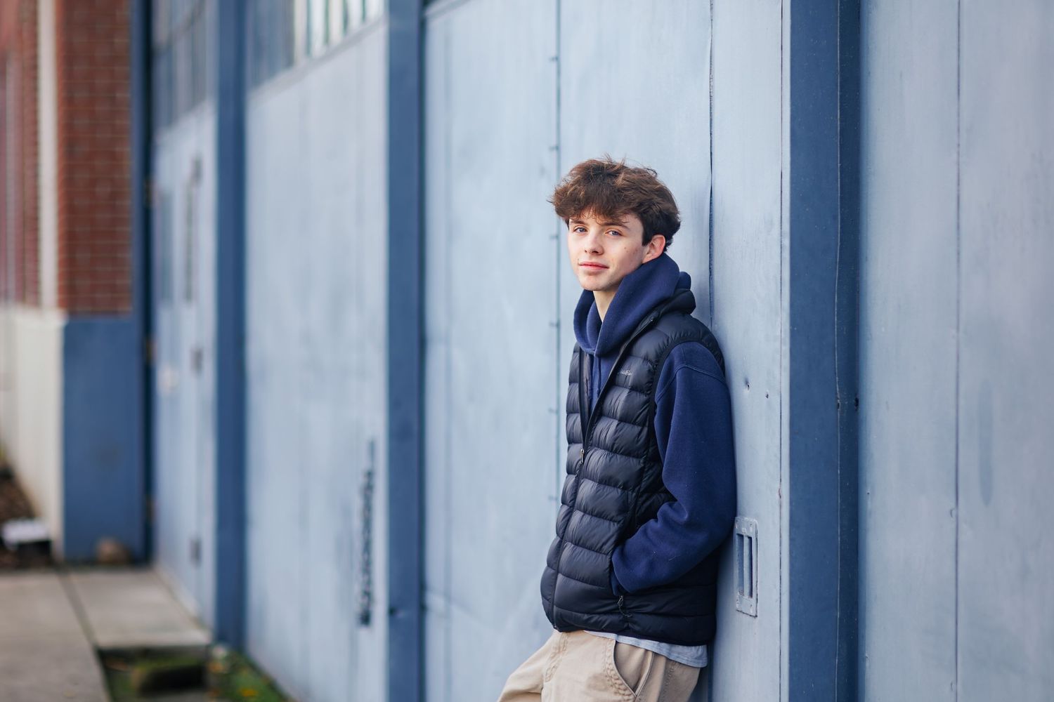 A teenaged boy in a navy blue puffer vest and hoodie leans casually against a blue-gray metal wall outside a building.