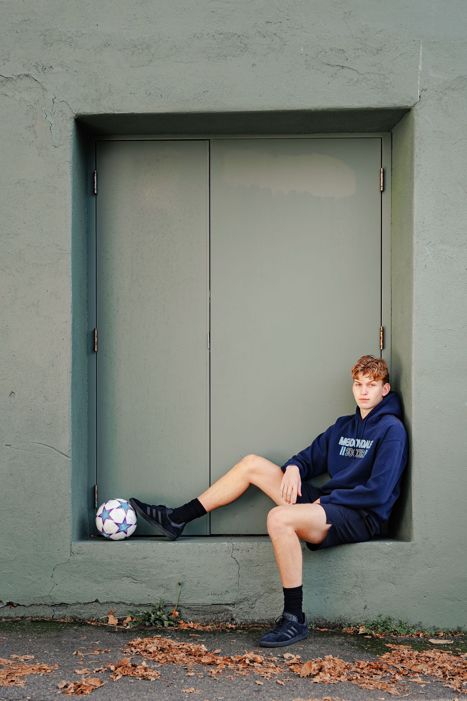 Soccer player resting in a concrete alcove with ball at feet wearing navy hoodie and shorts.