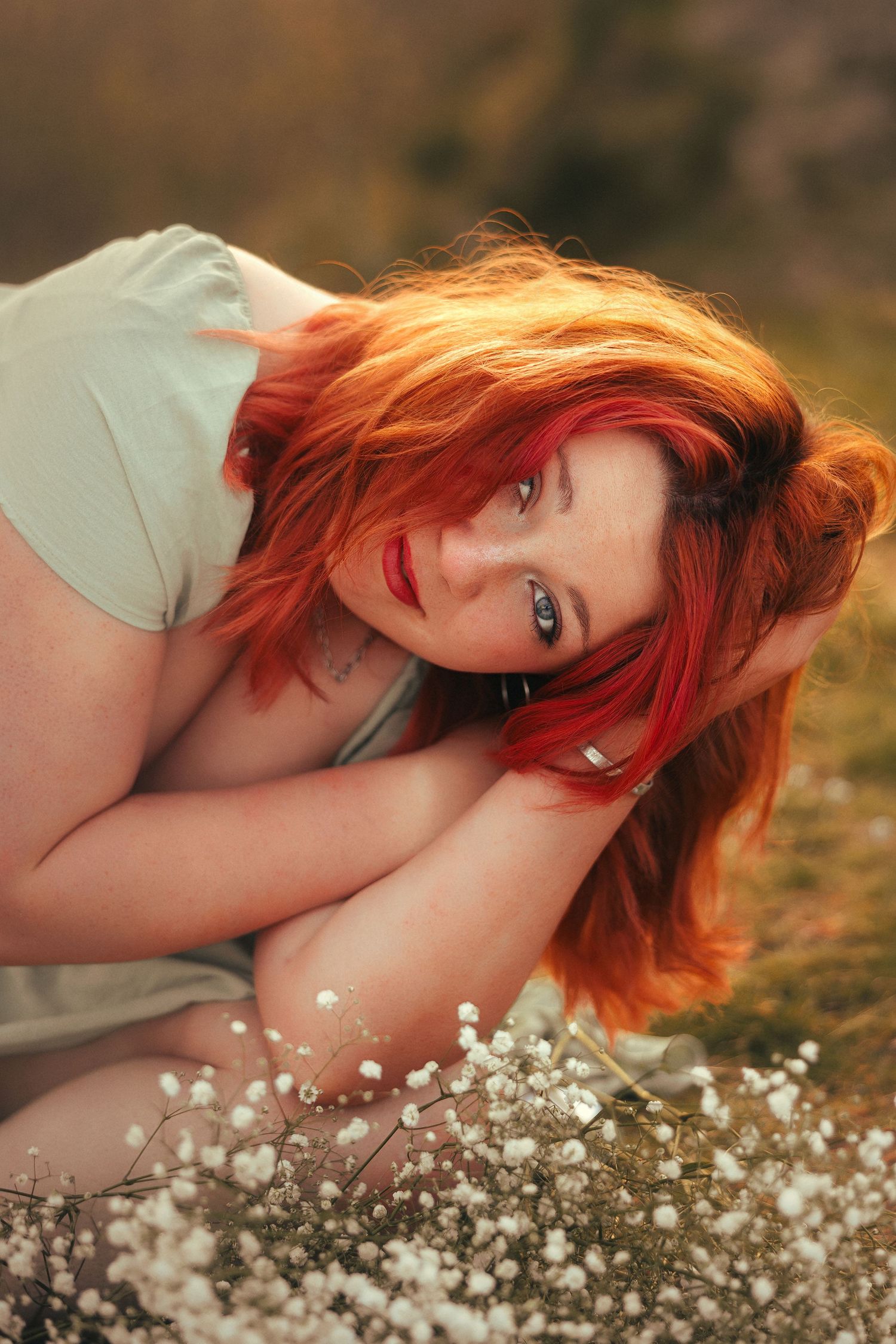 A dreamy portrait shows someone with vibrant red hair lying among delicate white baby's breath flowers in soft natural light.