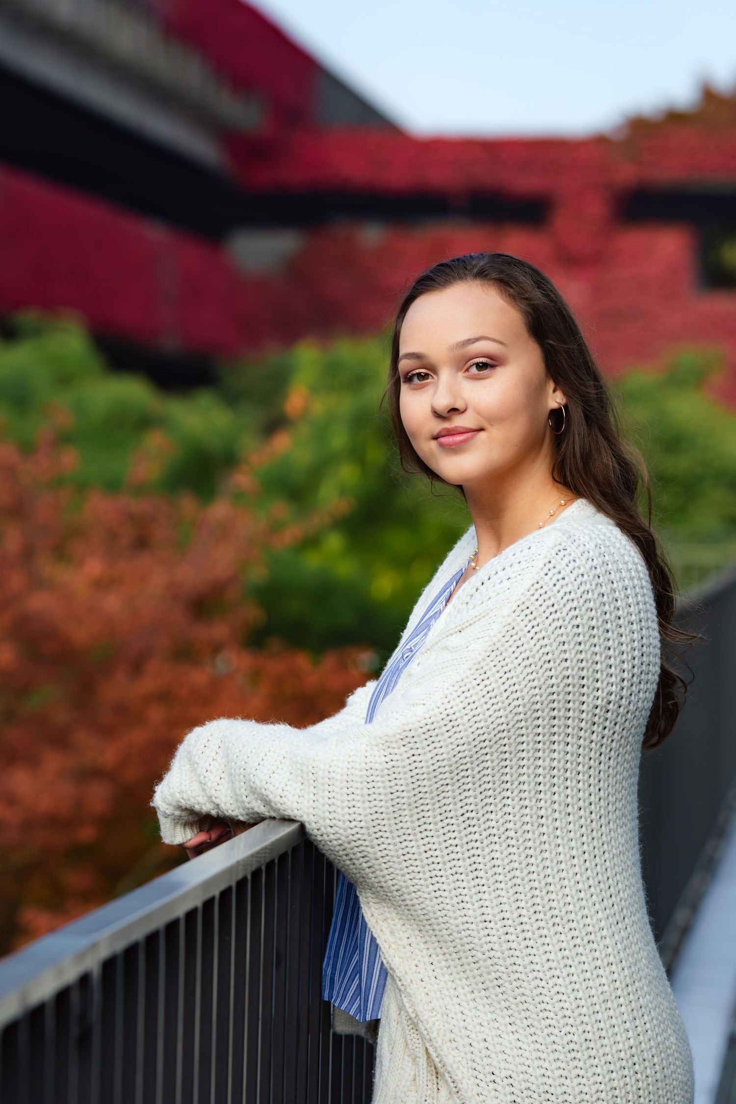 Woman in cream knit sweater leans on railing with red autumn foliage in background.
