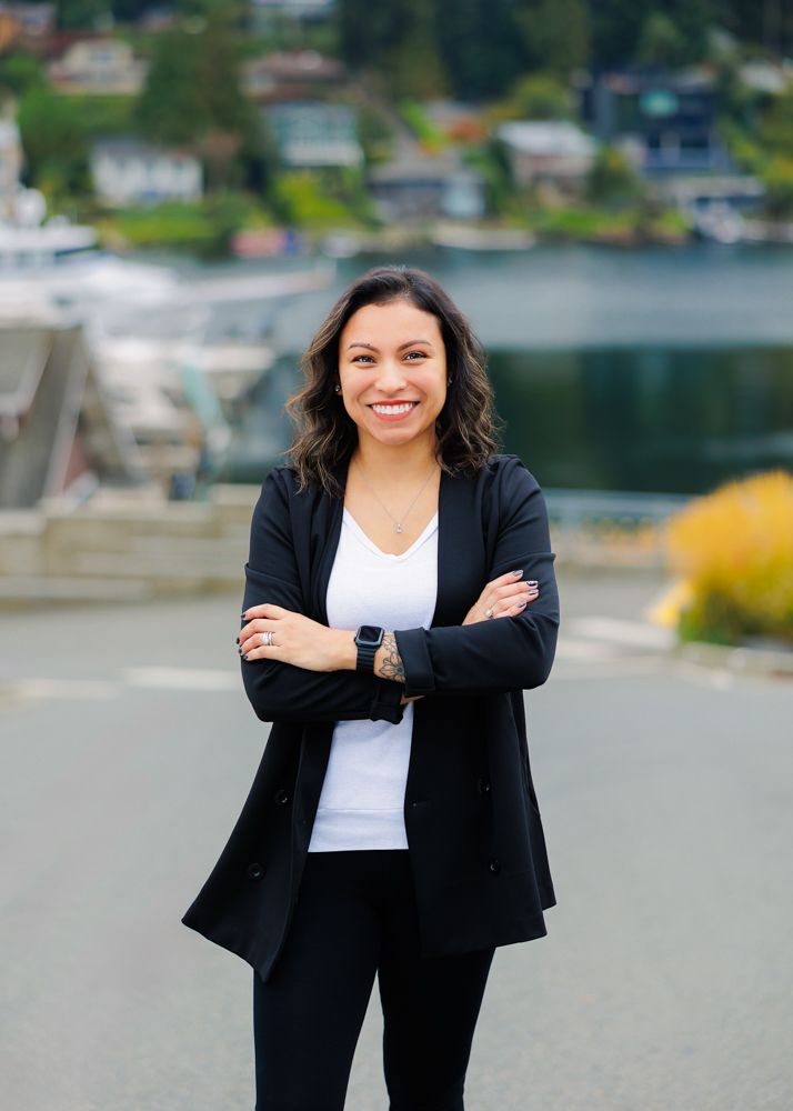 Professional woman wearing black blazer and white top stands in front of the waterfront with boats and houses in background.