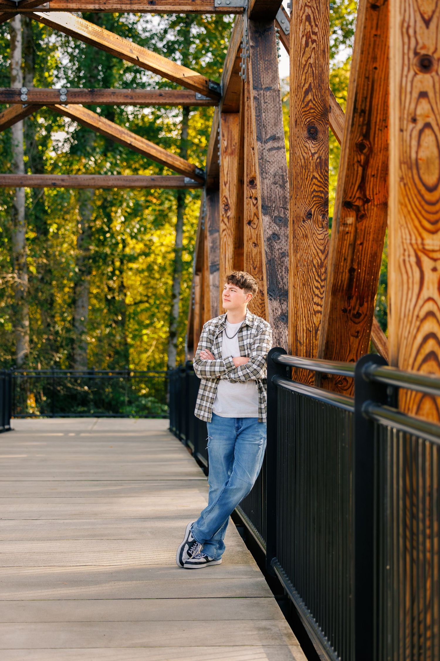 Person in jeans and plaid shirt walks along a wooden bridge railing with sunlit trees in the background.