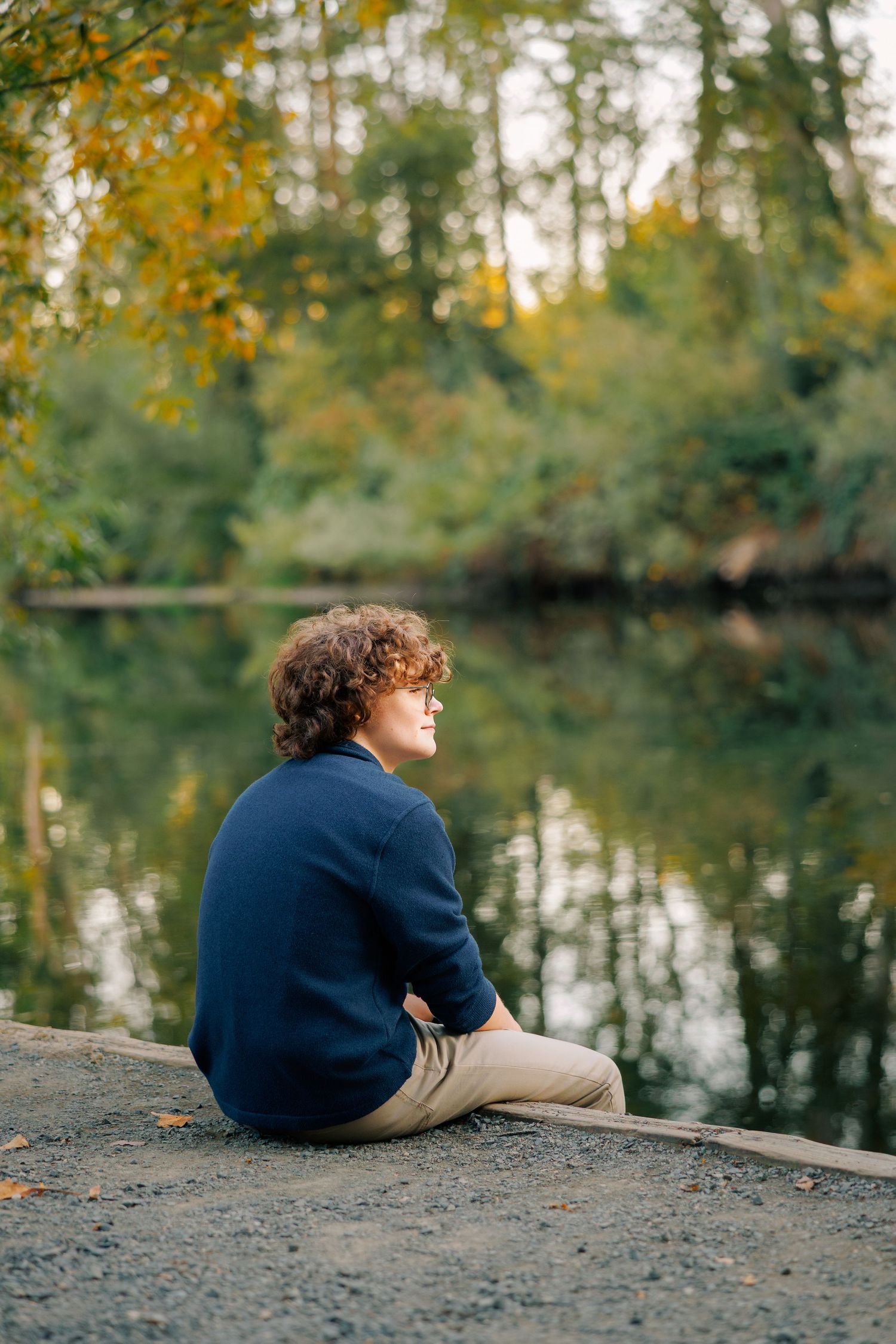 Person in navy sweater sits contemplatively by a tranquil pond surrounded by autumn trees reflecting in the water.
