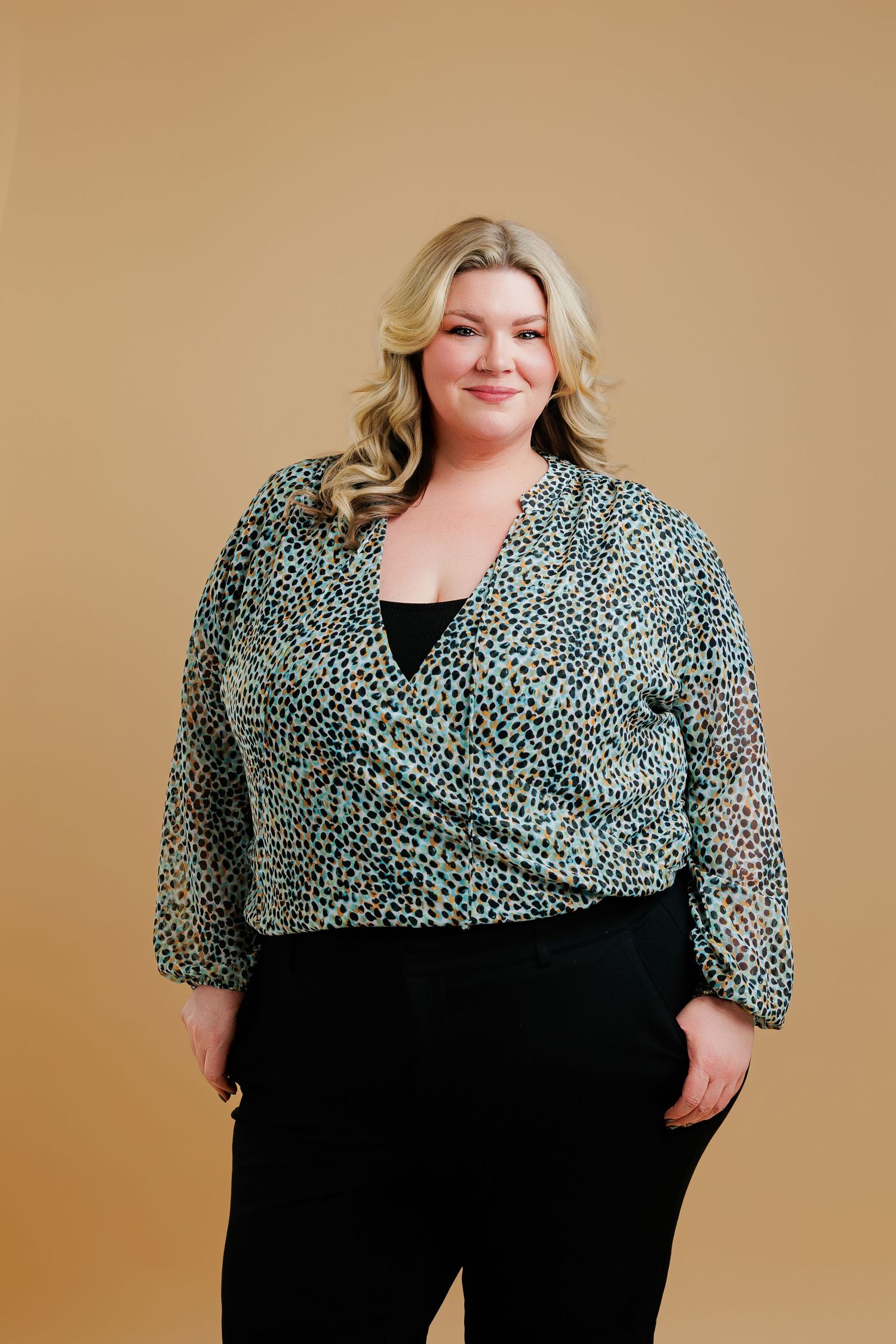 Plus size model wearing a black and white patterned wrap top with black pants against a beige background.
