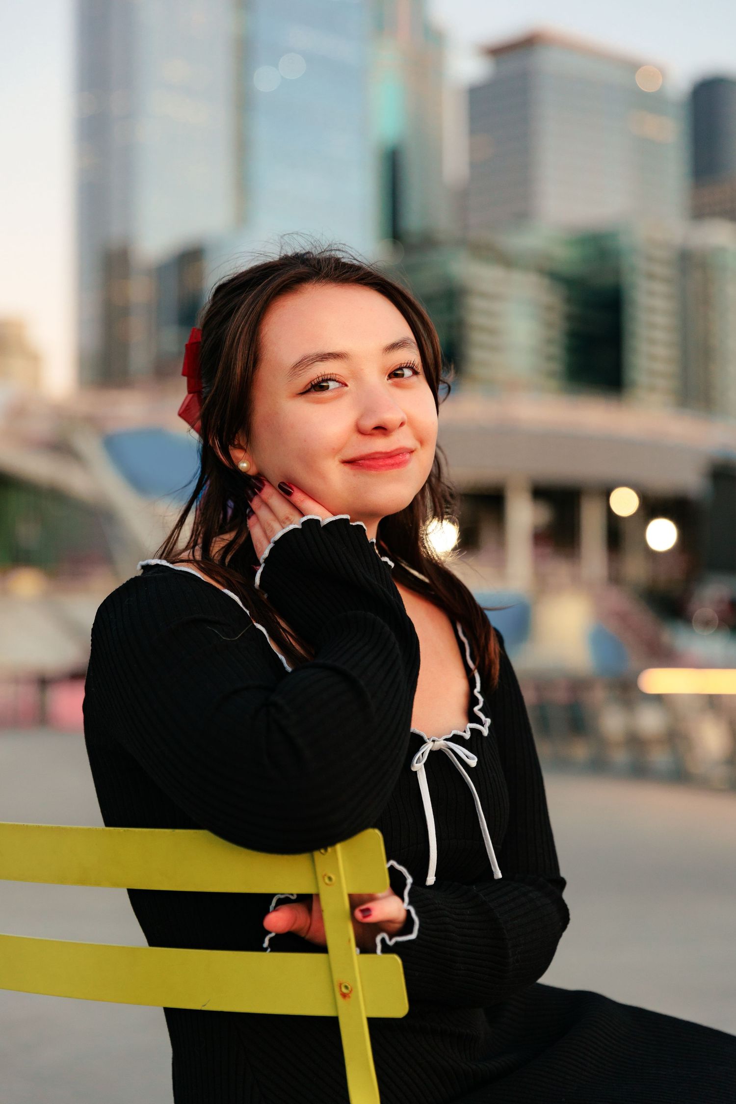Portrait of someone in a black sweater sitting on a yellow chair against an urban cityscape background at dusk.