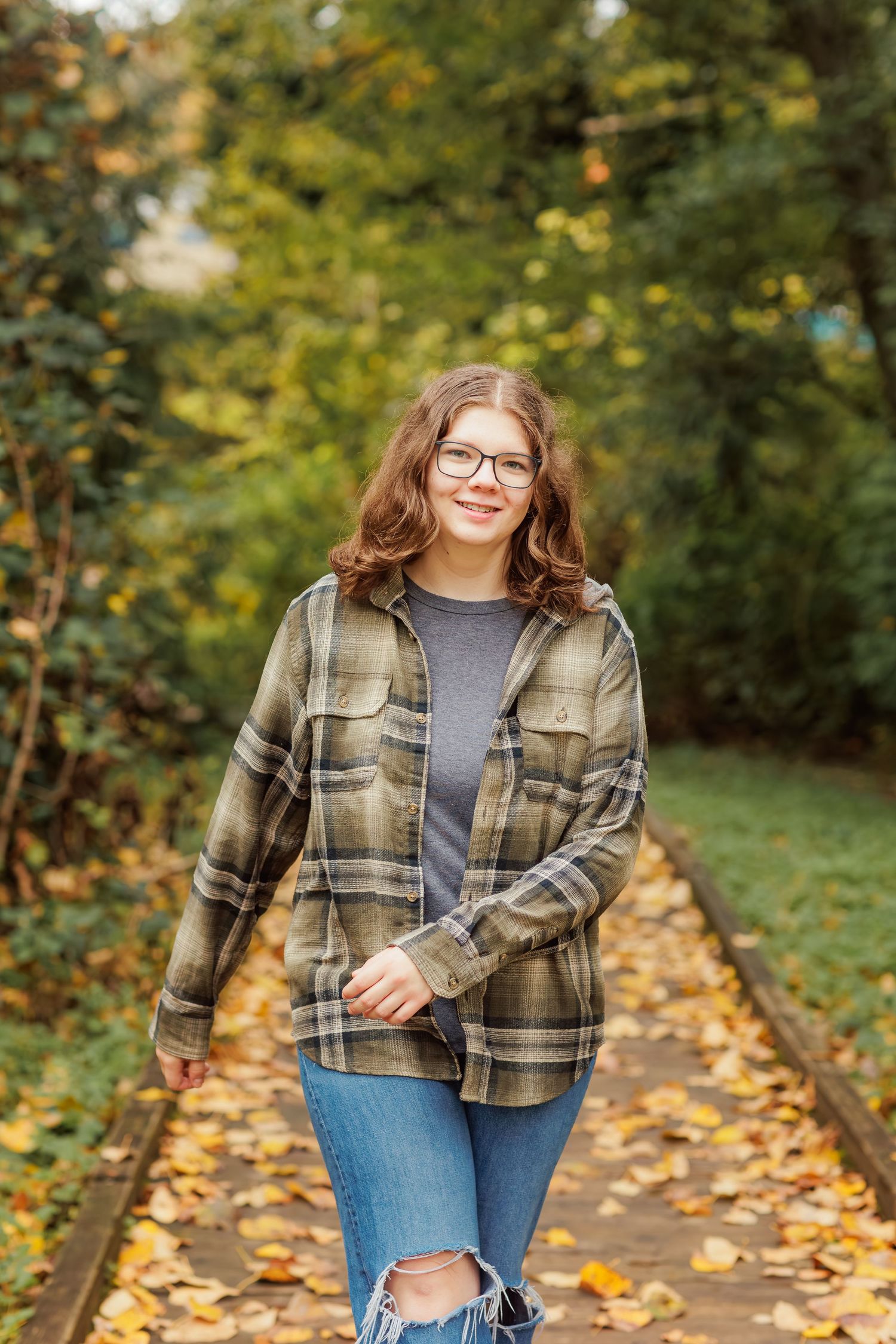 A teen girl in a plaid flannel jacket and jeans walks down a wooden boardwalk in autumn in the park.