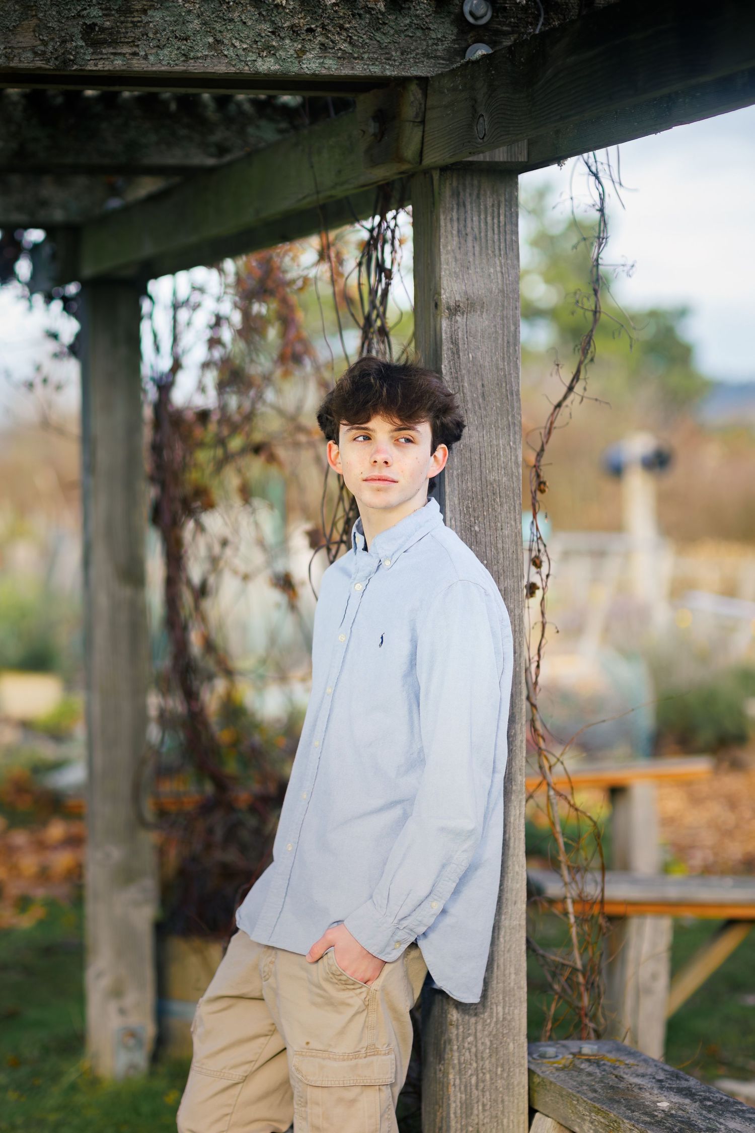 A teen boy in a light blue button-down shirt and khaki pants poses against wooden posts in an autumn outdoor setting.