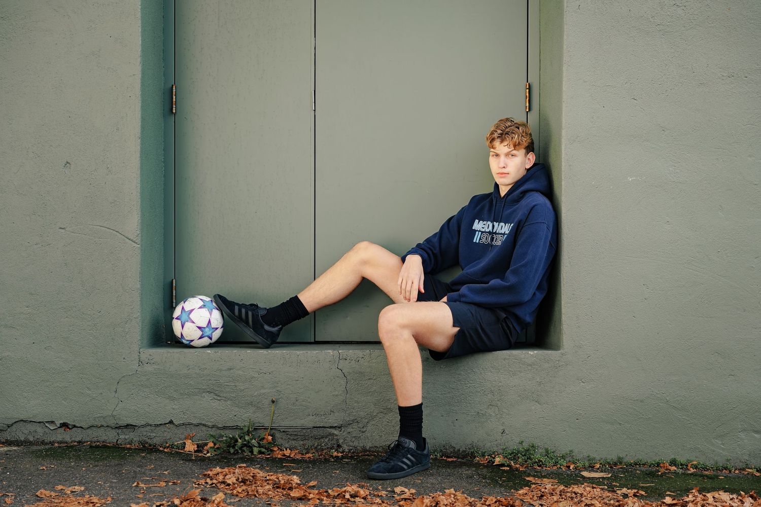 A high school boy in a navy sweatshirt and shorts sits in a wall alcove with his soccer ball at his feet.