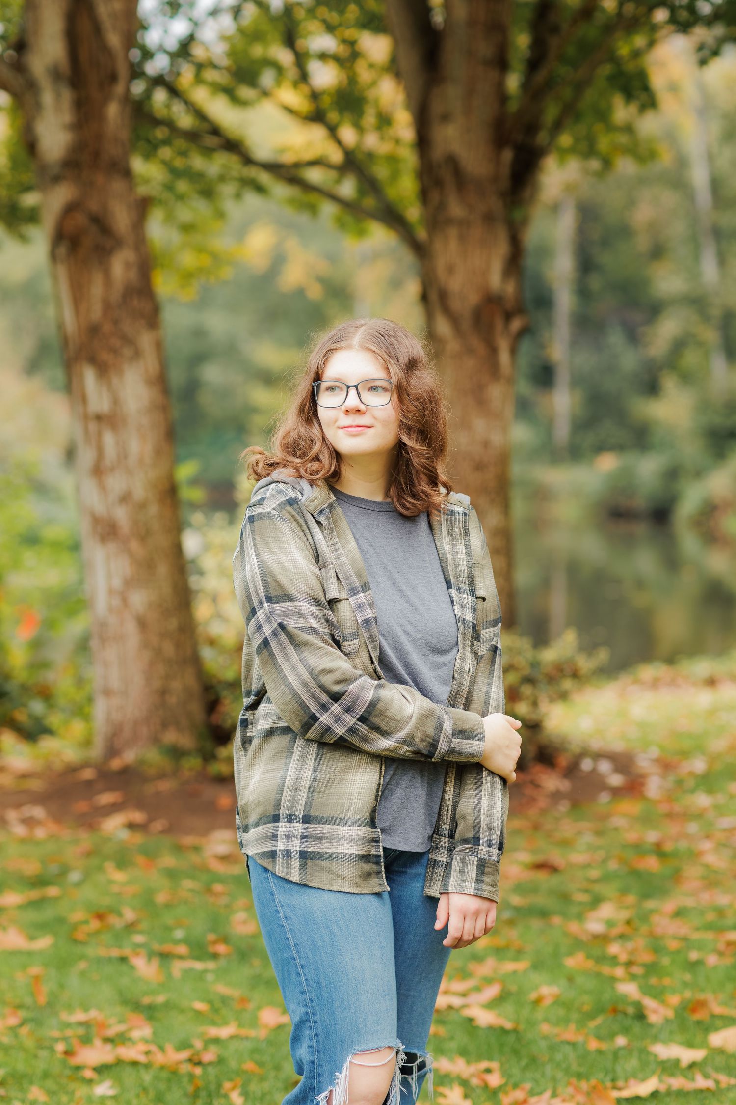 A person in a plaid flannel jacket and jeans poses casually against autumn trees with fallen leaves on the ground.