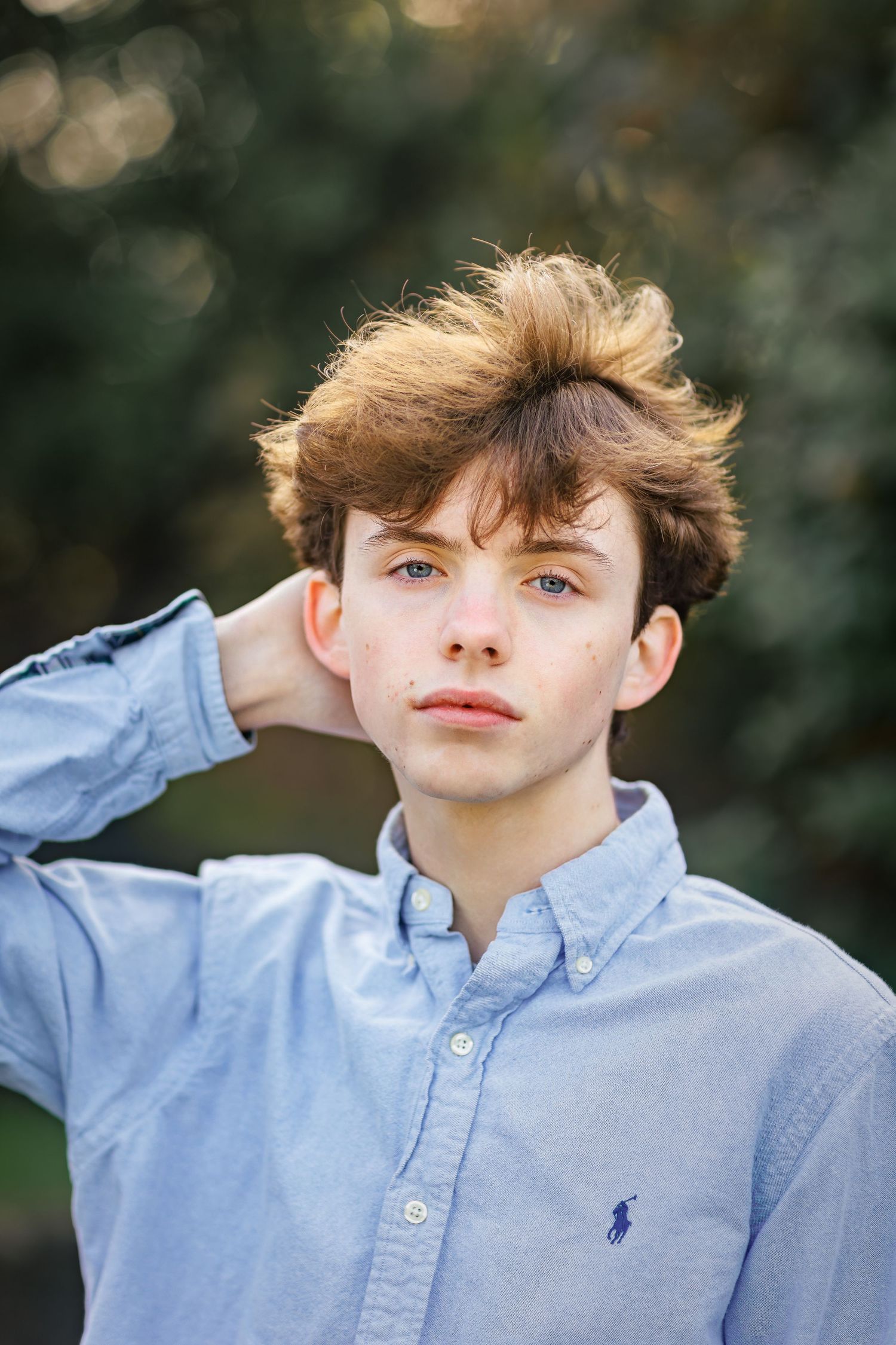 Young person in light blue Ralph Lauren button-down shirt poses outdoors with tousled brown hair against blurred background.