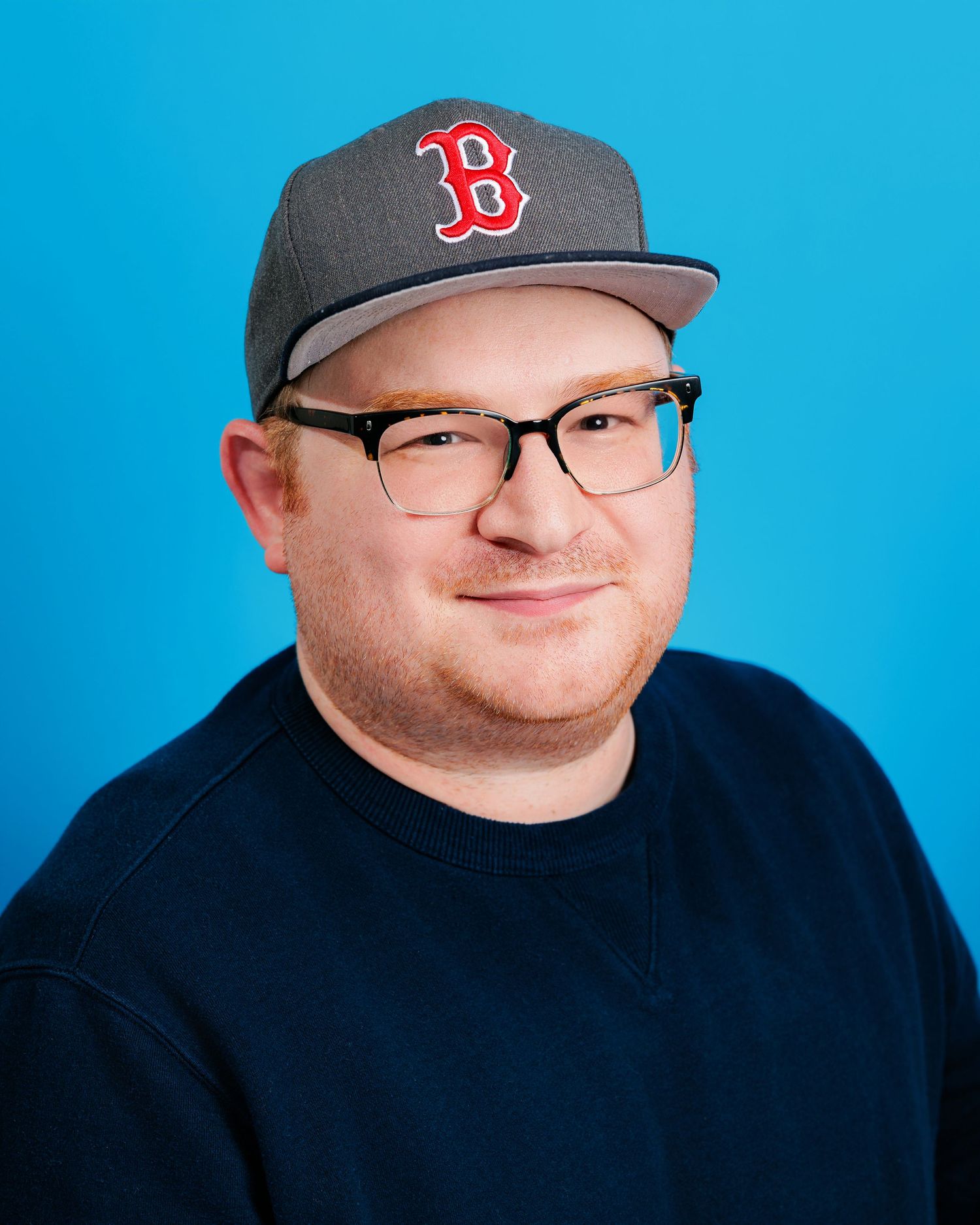 A smiling figure wearing a Boston Red Sox baseball cap and dark shirt against a bright blue background.