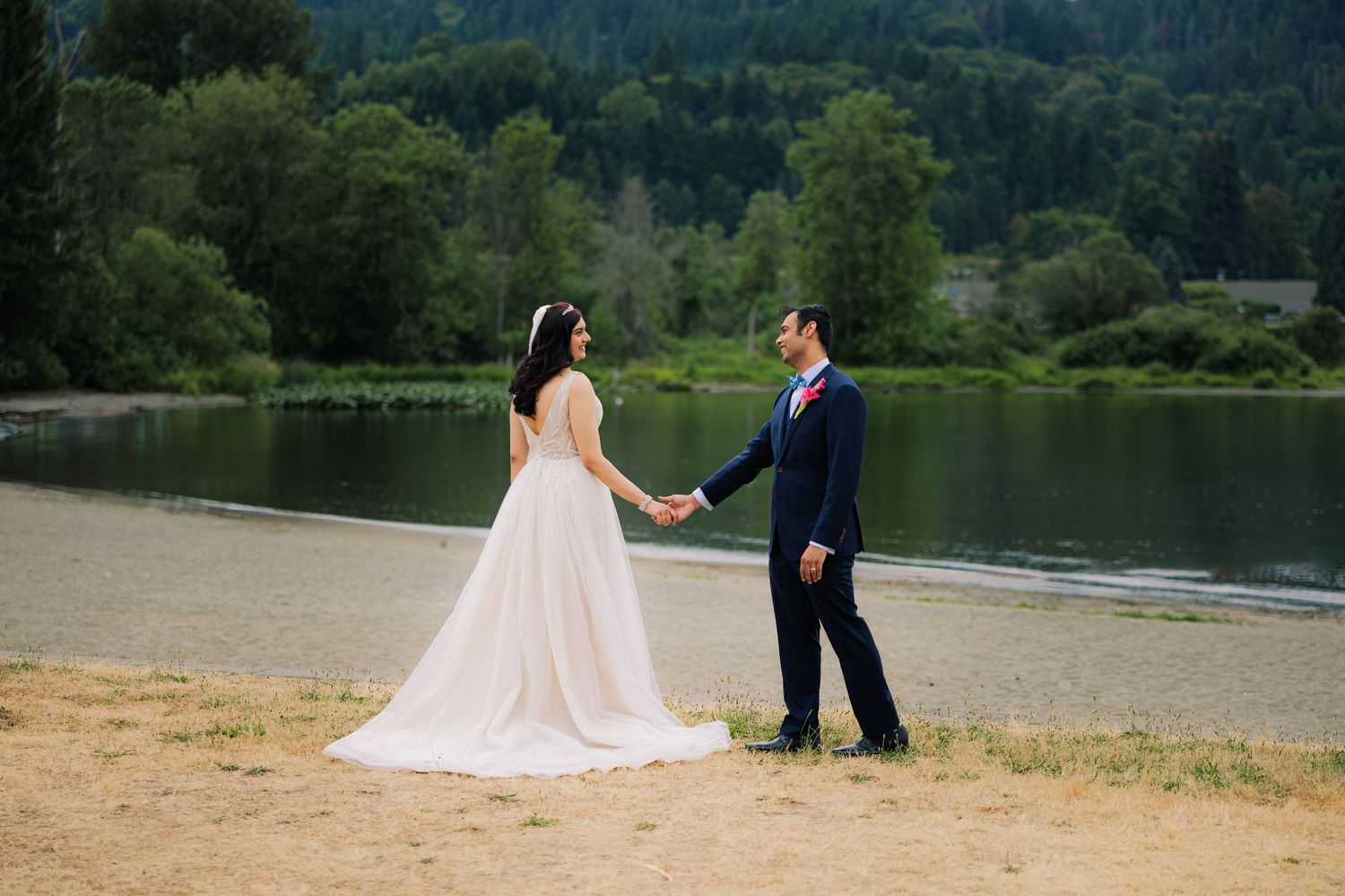 A romantic wedding couple holds hands by a serene lake with mountains and forest in the background.